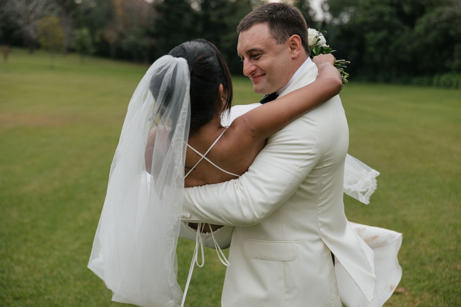 A joyful bride and groom embrace on a green lawn, the groom smiling in a white suit as the bride wears a flowing veil.