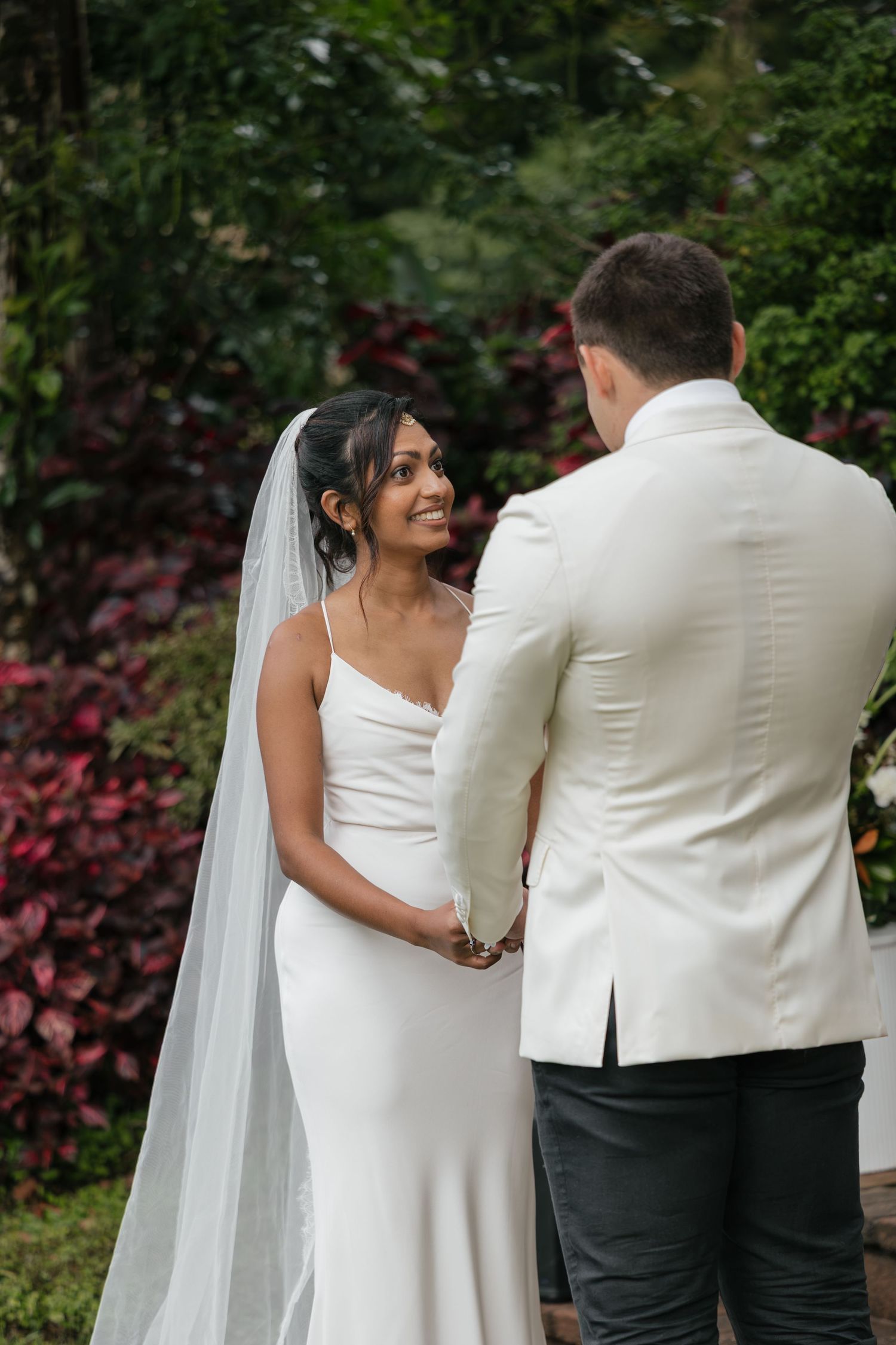 Bride in white gown and veil holds hands with groom in white jacket during outdoor garden wedding ceremony.