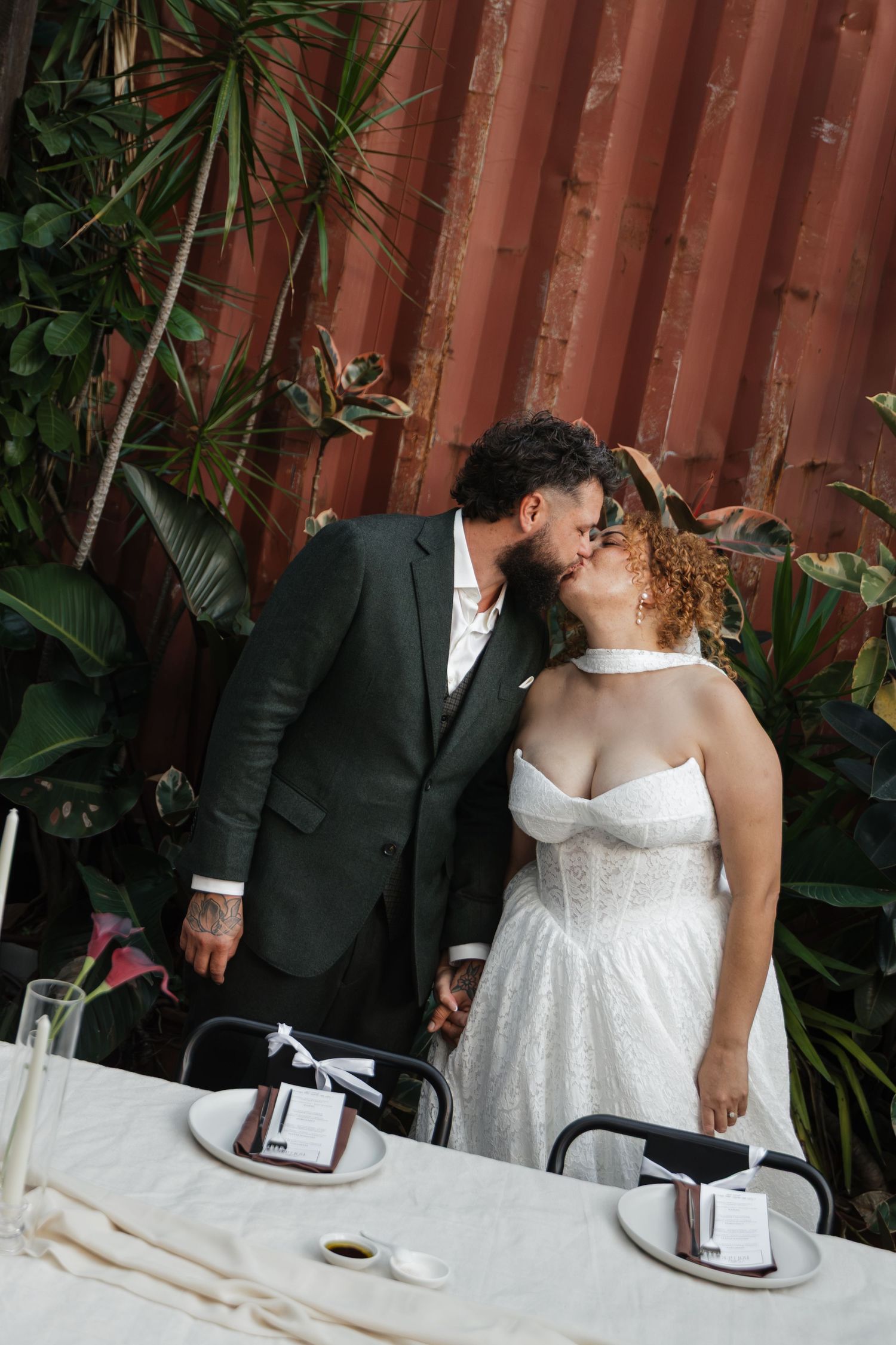A couple in wedding attire shares a kiss at a decorated table surrounded by tropical plants and a rustic metal wall backdrop.