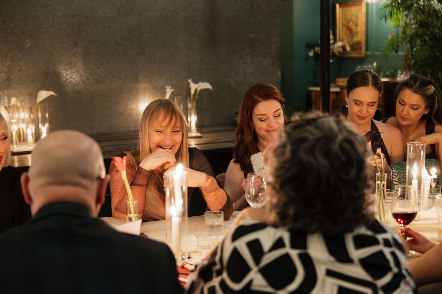 Women laughing and socializing at a candlelit dinner party in a cozy restaurant setting with wine glasses.