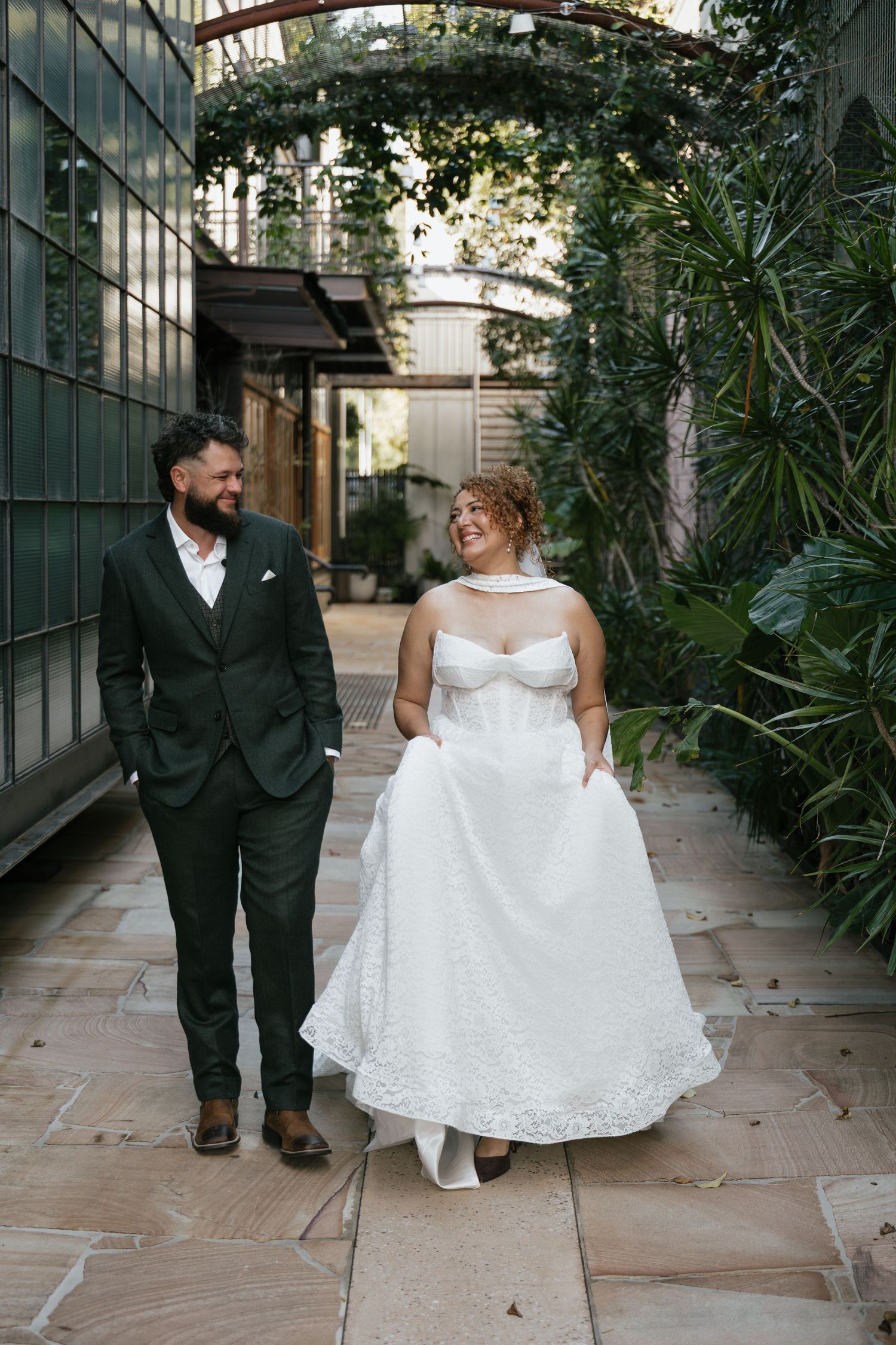 A bride and groom walk together laughing in a lush garden courtyard, she lifts her white lace gown as he wears a dark green suit.