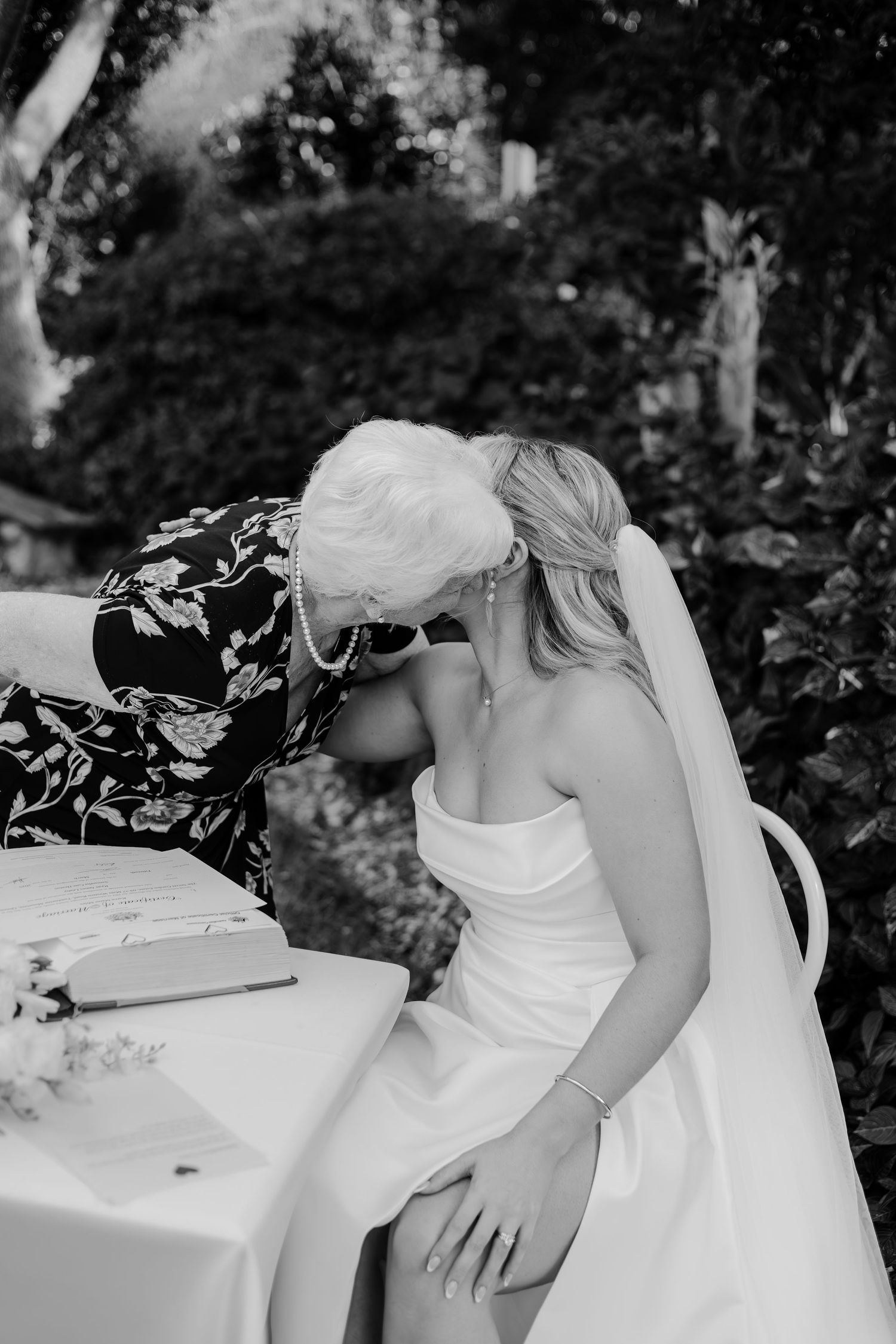 Black and white photo of a bride in strapless gown sharing an emotional moment with an older woman at an outdoor wedding.