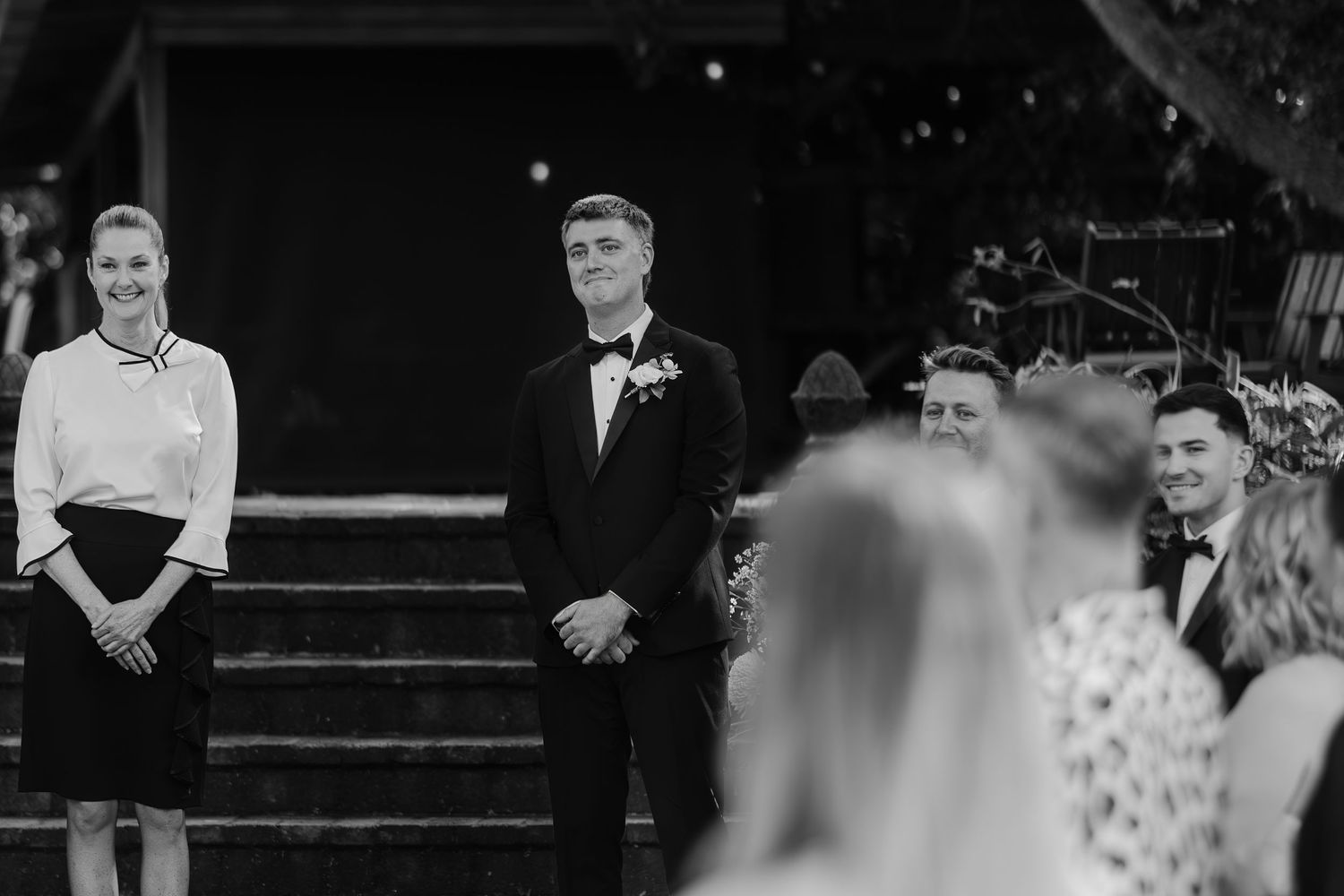 Groom in black tuxedo smiling at altar during outdoor wedding ceremony with guests in foreground.