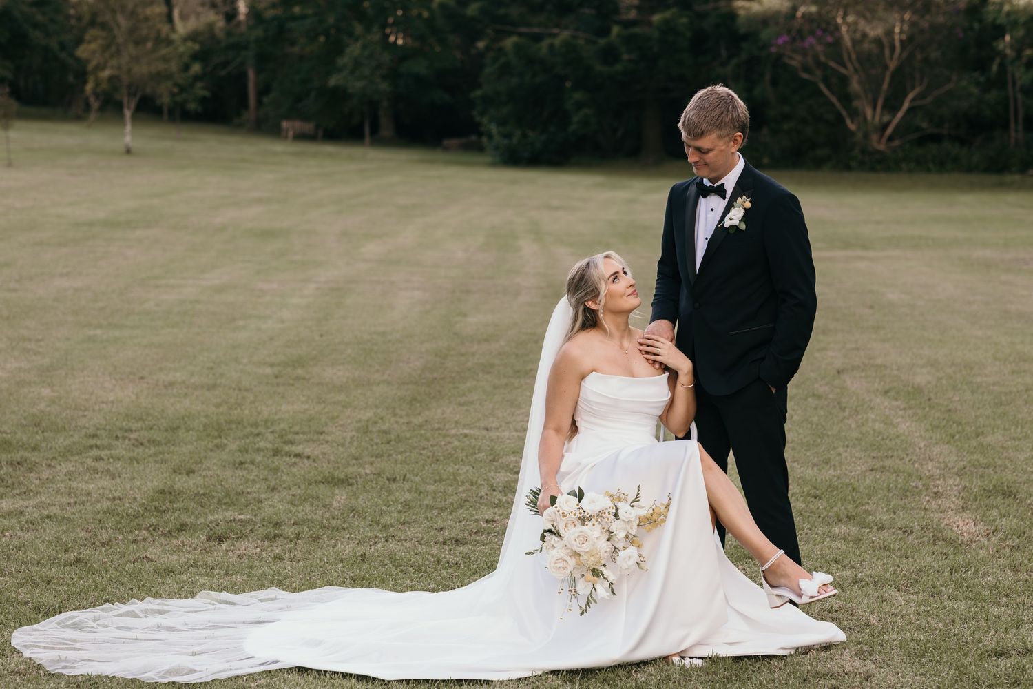 Bride in white gown sits on grass holding bouquet as groom in black tuxedo stands beside her outdoors.