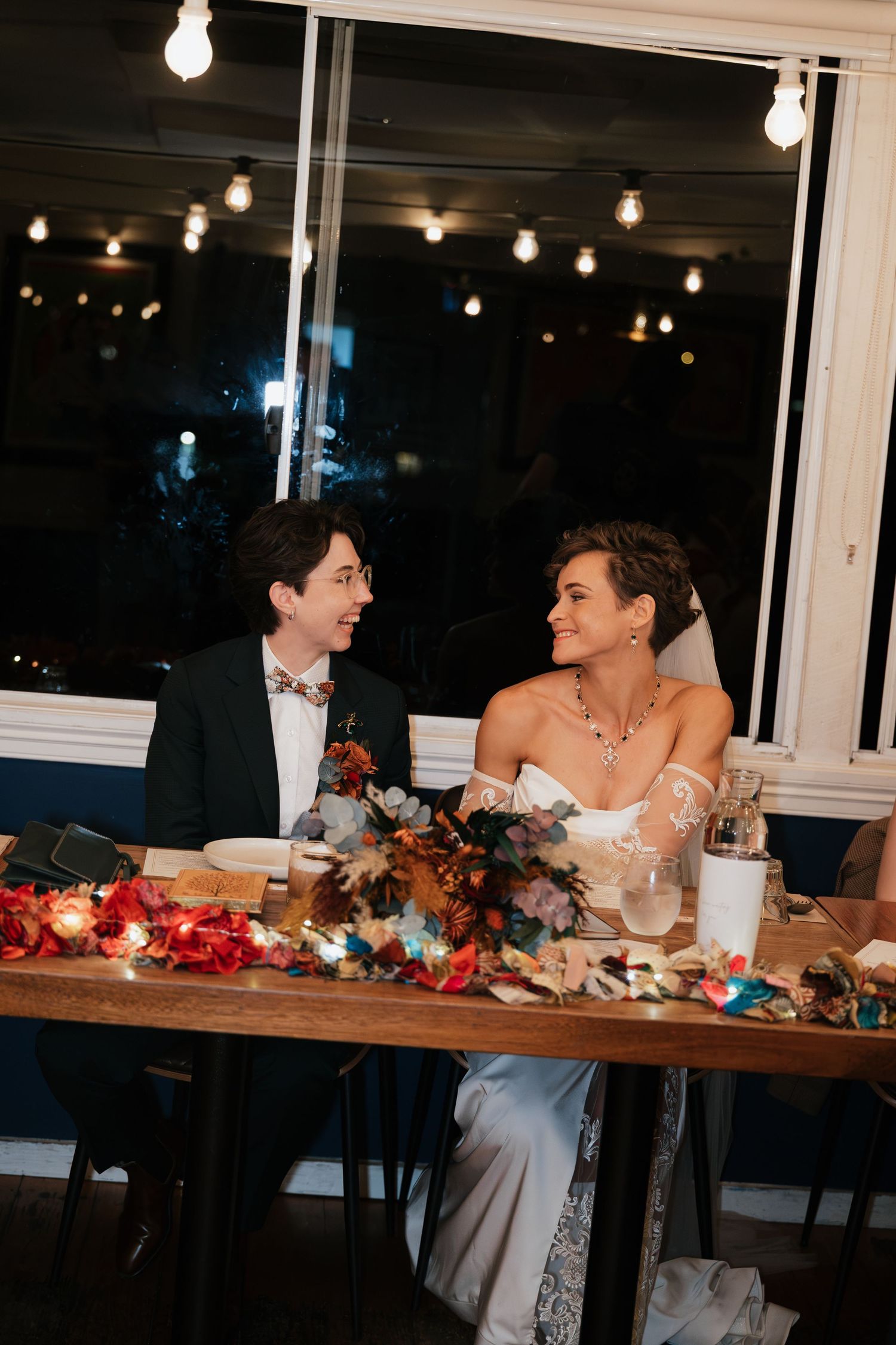 Two brides share a joyful moment at their wedding reception table adorned with colorful floral arrangements and string lights.