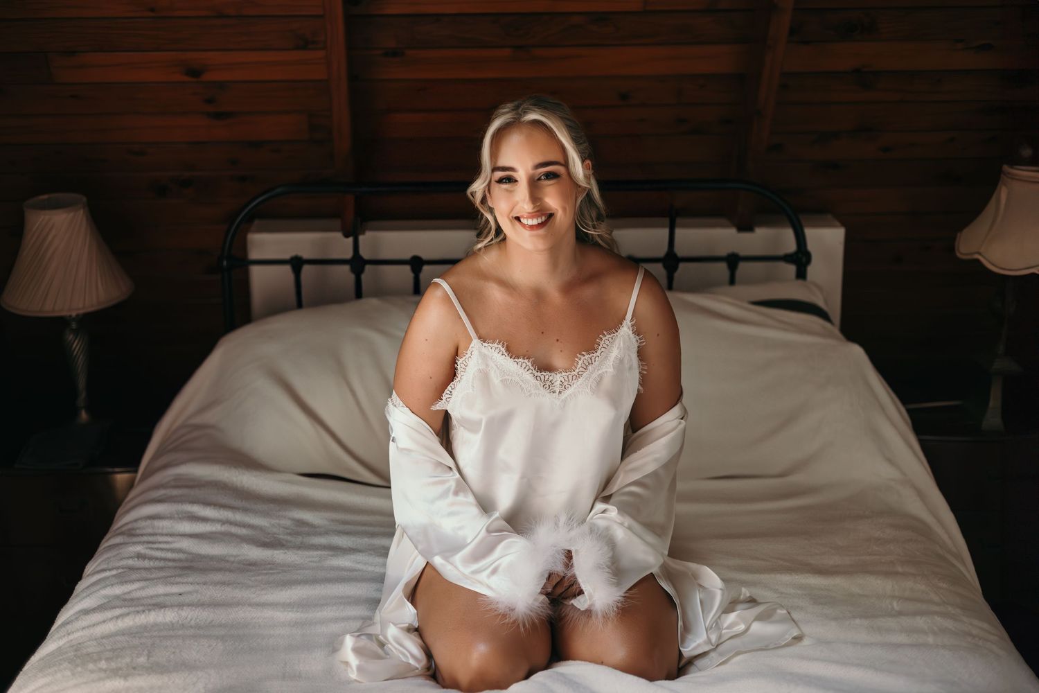 A smiling blonde woman in white satin pajamas sits on a bed in a rustic wooden bedroom with warm lamp lighting.