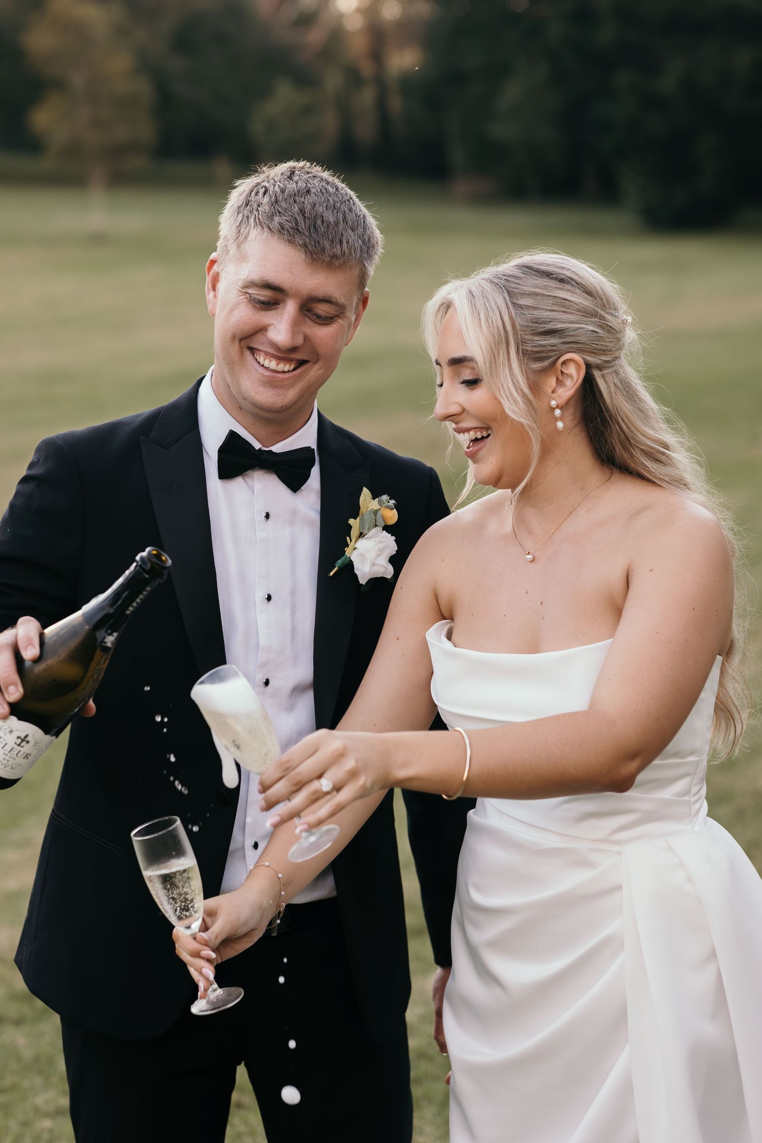 Bride and groom smiling while pouring champagne outdoors, groom in black tuxedo, bride in strapless white gown.