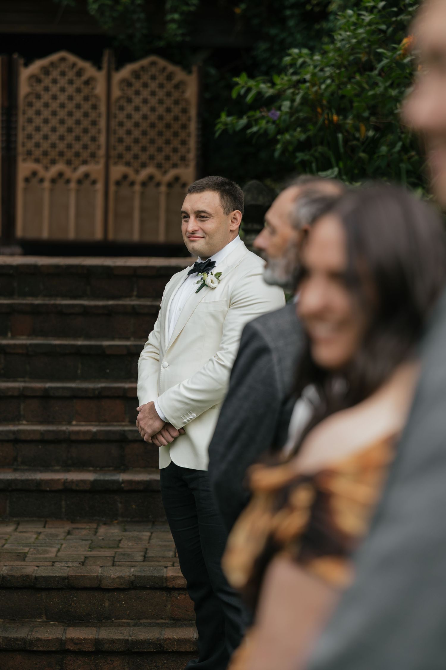 Groom in white tuxedo jacket and bow tie smiling at wedding ceremony on brick steps surrounded by guests.