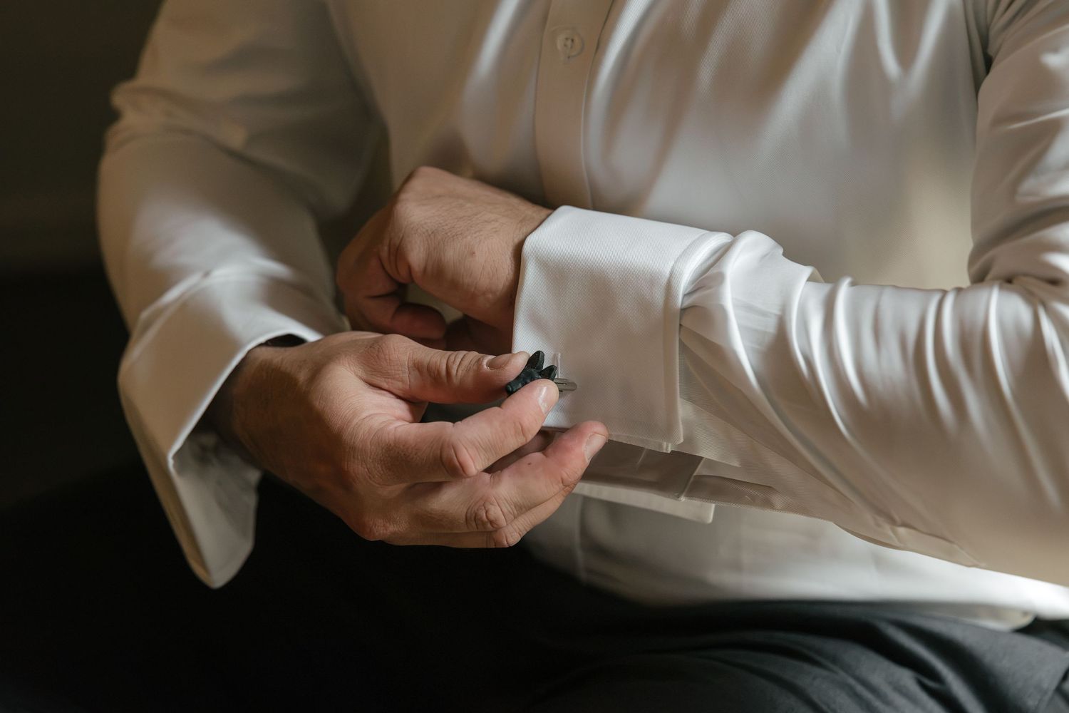 Man in white dress shirt fastening dark cufflinks, close-up detail shot for formal wedding attire.