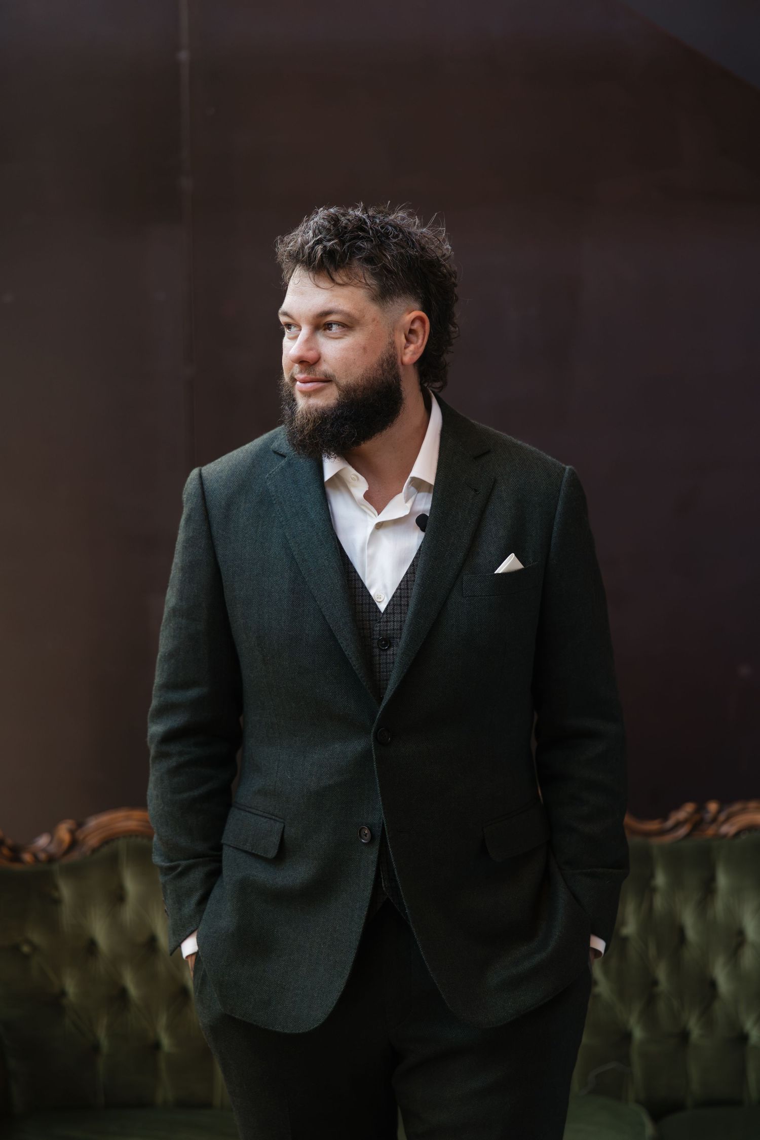 Bearded man in dark green suit with white pocket square standing confidently in an elegant interior setting.