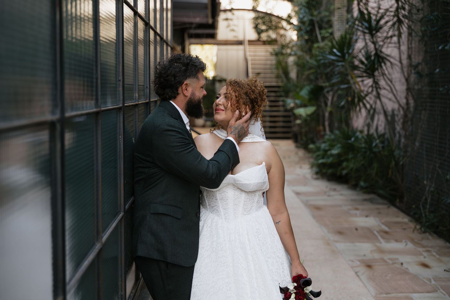 Bride and groom share a romantic moment in an outdoor alley, with the bride in a white gown holding red flowers.