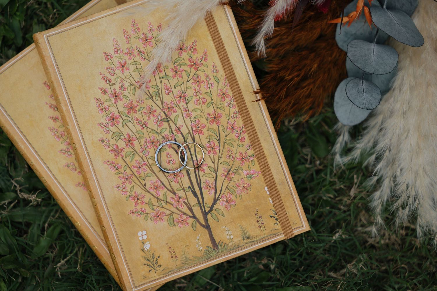 Two floral tree wedding invitations with gold rings on grass, featuring pink blossoms on yellow background.