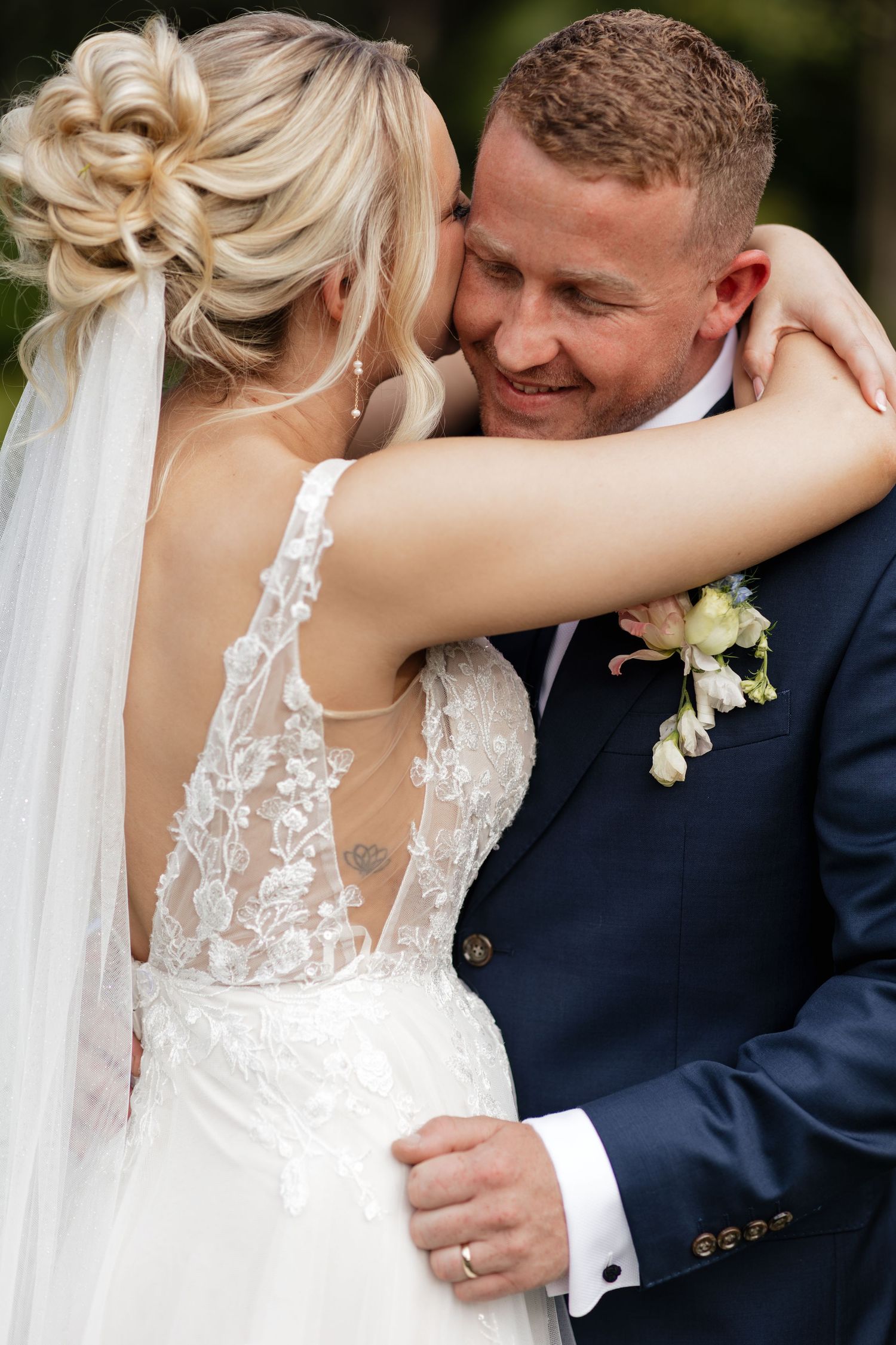 Bride in lace wedding dress embraces smiling groom in navy suit with boutonniere during outdoor wedding ceremony.