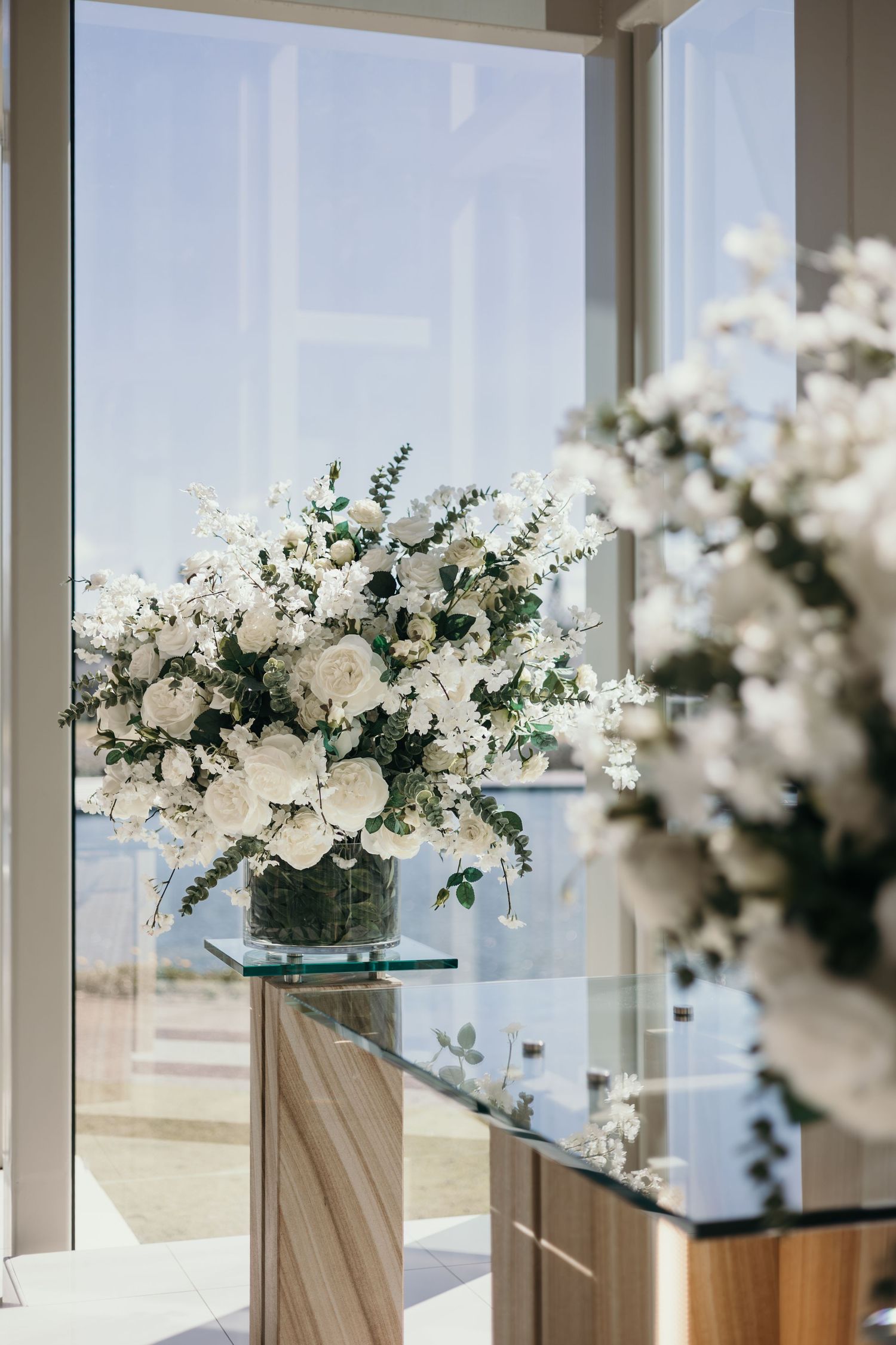 Elegant white floral arrangements in glass vases line a mirrored surface with natural light streaming through windows.