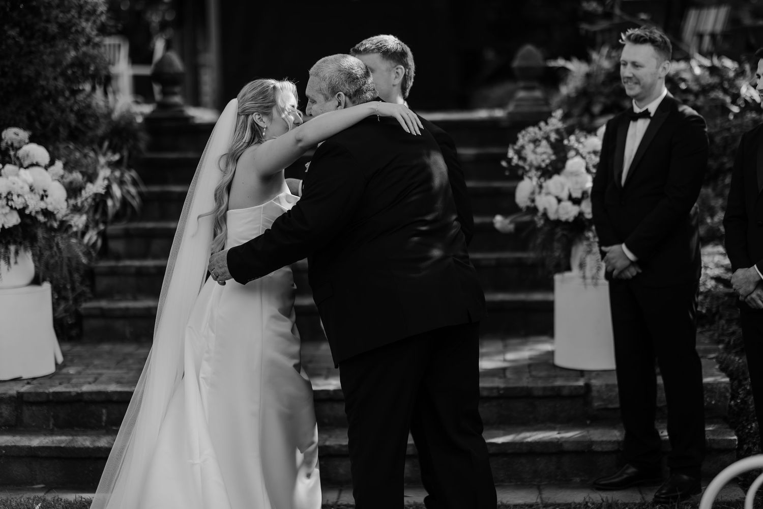 Bride hugging her father at outdoor wedding ceremony while groom watches, black and white wedding photo.