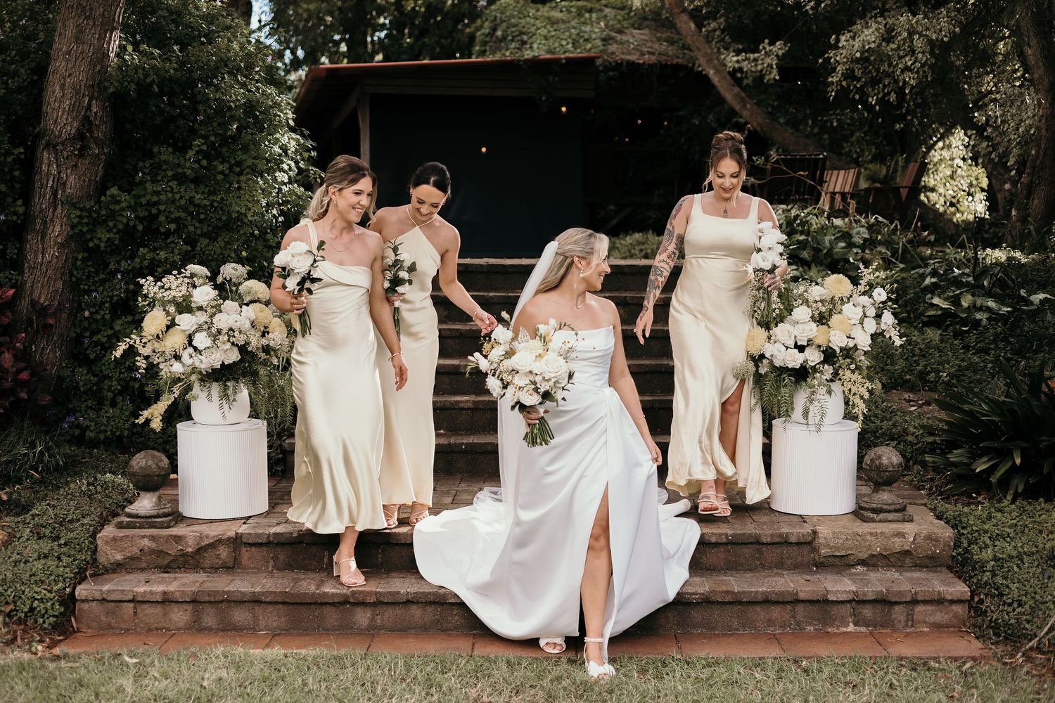 Bride in white gown descends stone steps with three bridesmaids in gold dresses, surrounded by white floral arrangements.