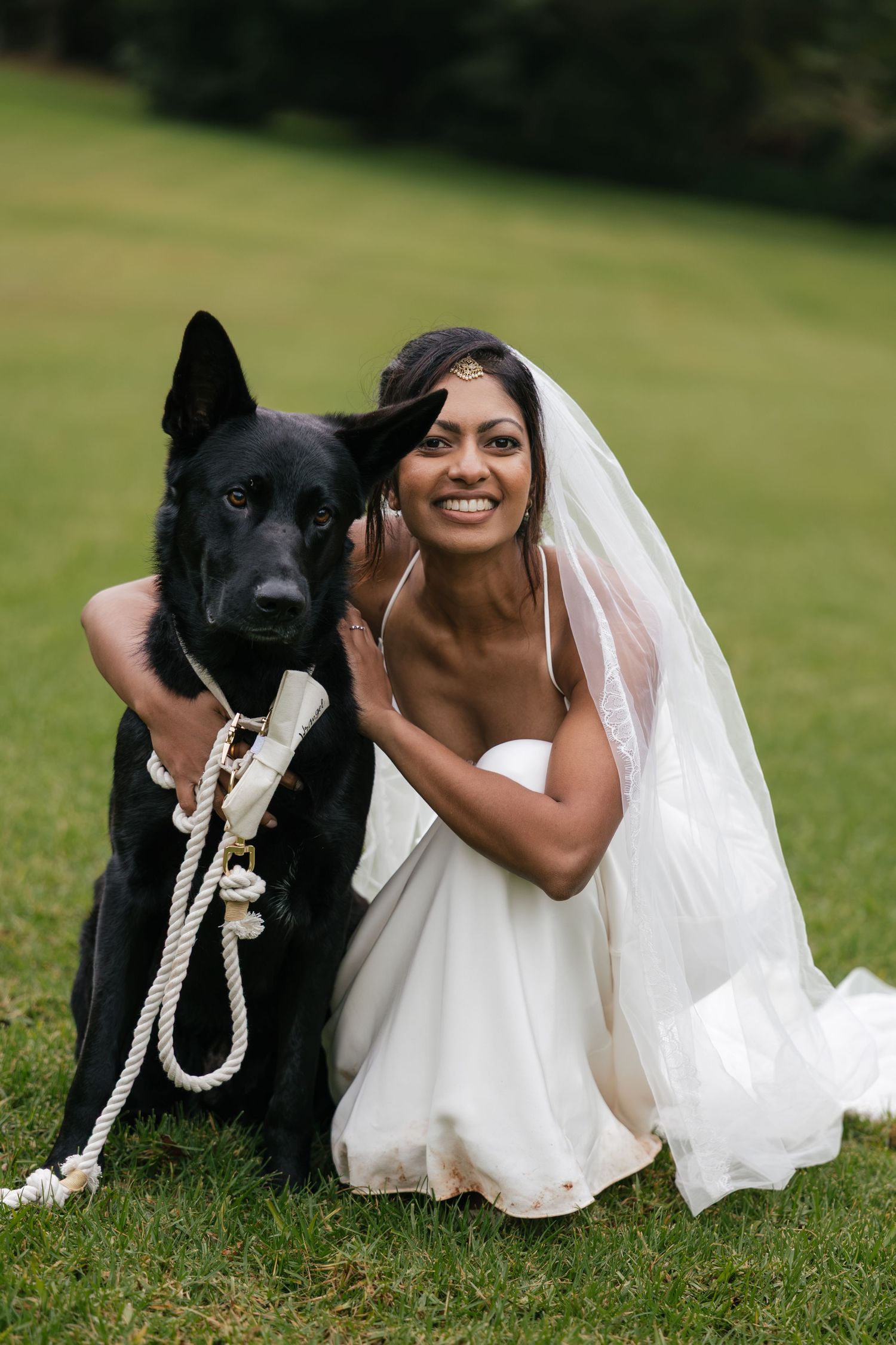 Smiling bride in white gown and veil kneeling beside a black German Shepherd on green grass.