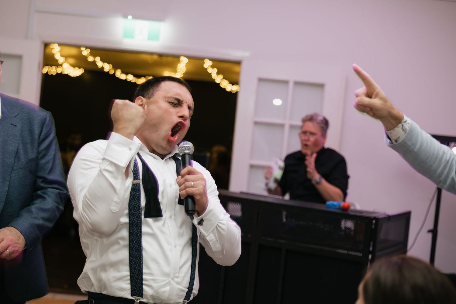 A man in suspenders passionately sings into a microphone at a wedding reception while guests celebrate in the background.