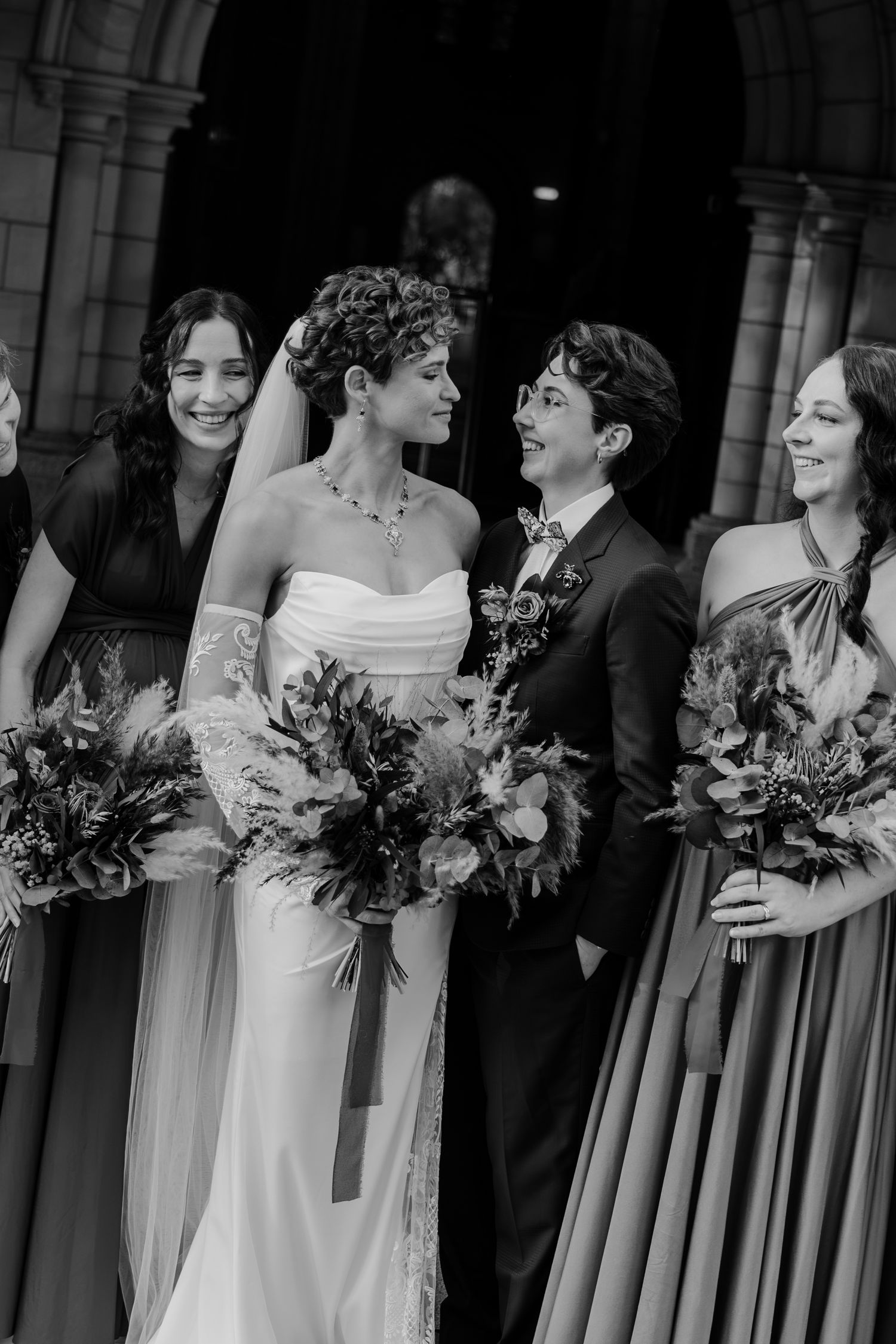 Black and white wedding photo of bride in white gown with partner in suit, flanked by bridesmaids holding bouquets.