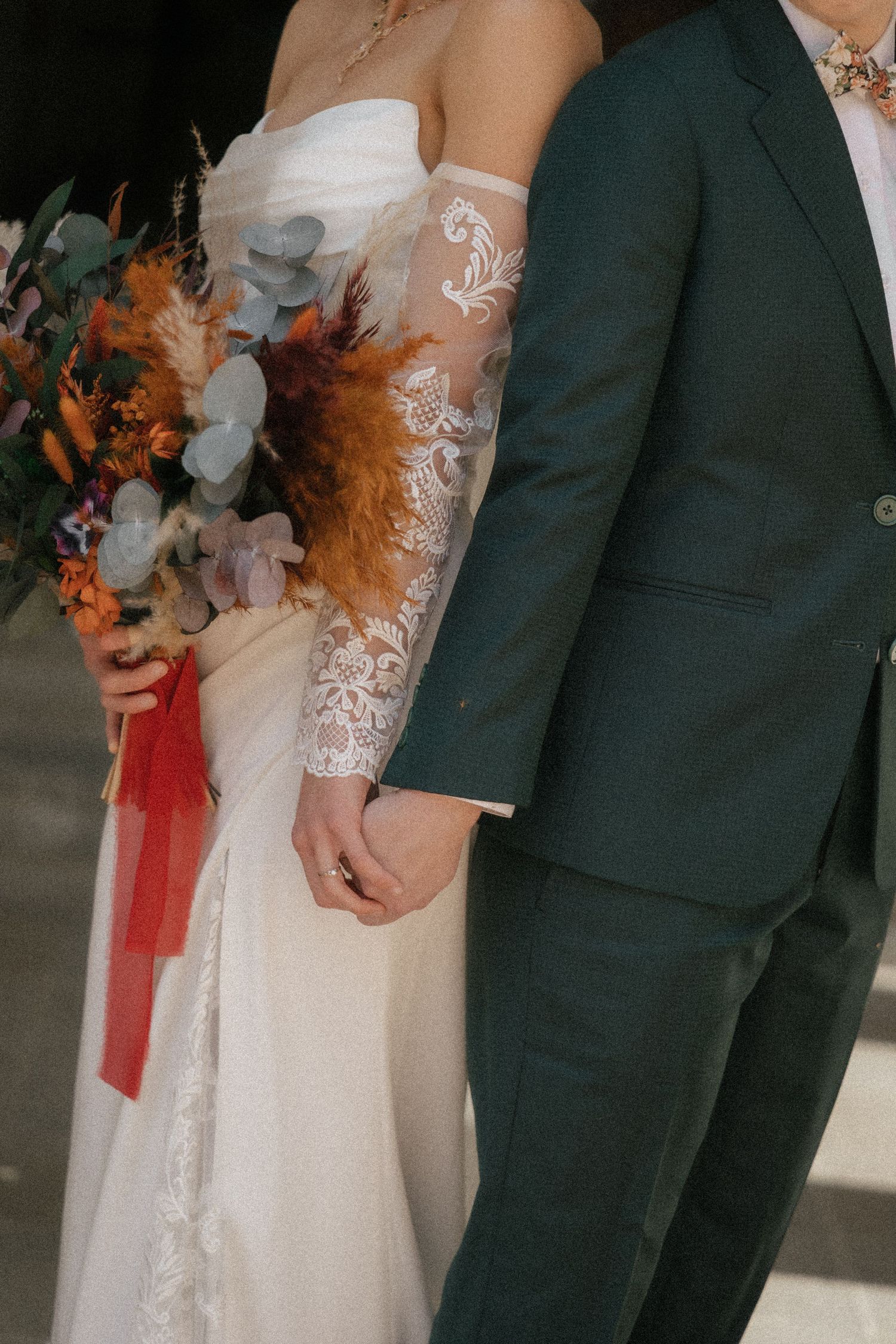 Bride in lace gloves holding colorful bouquet with dried flowers stands beside groom in dark green suit.