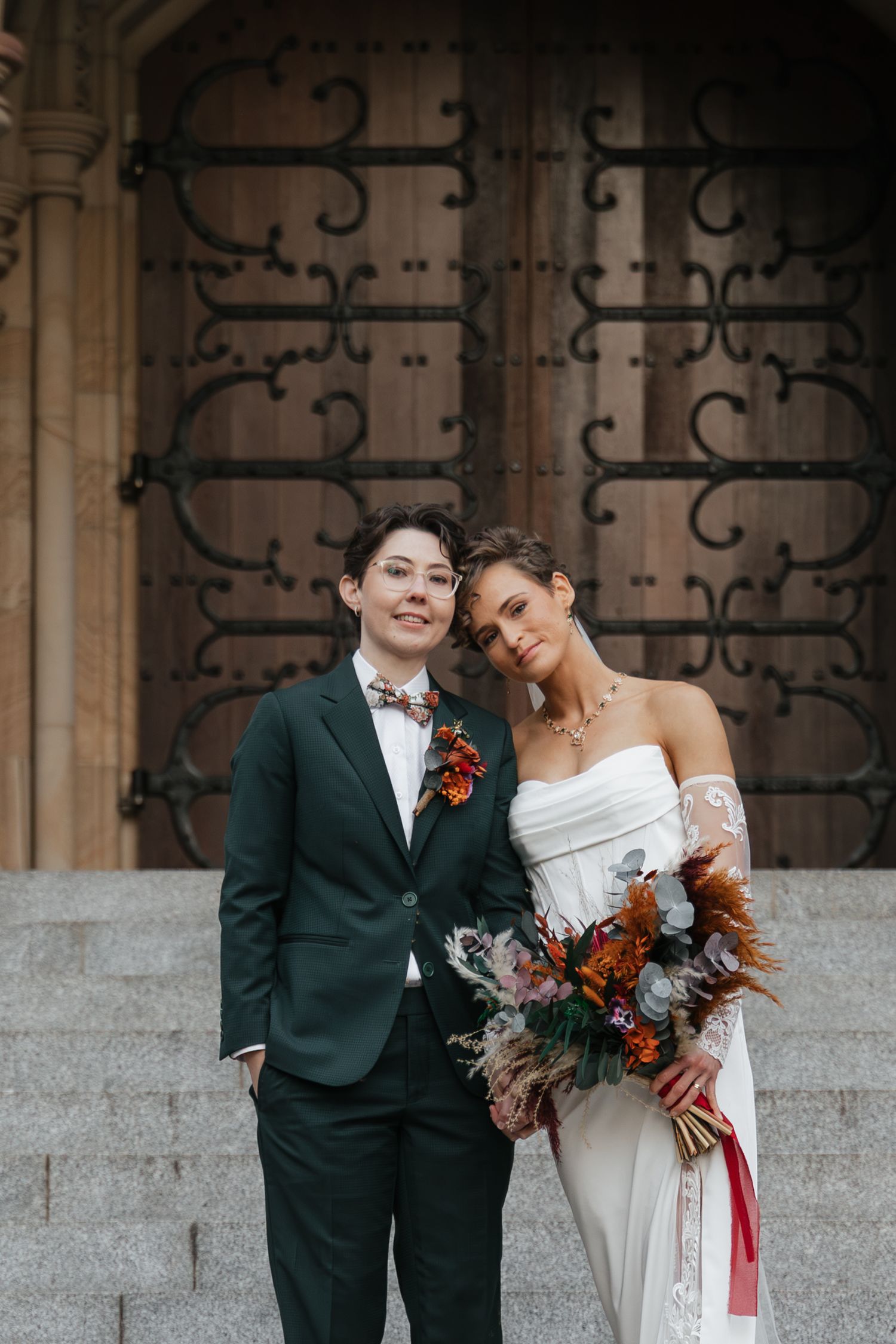 Two brides pose on church steps, one in a dark suit with boutonniere, one in a white gown holding autumn bouquet.