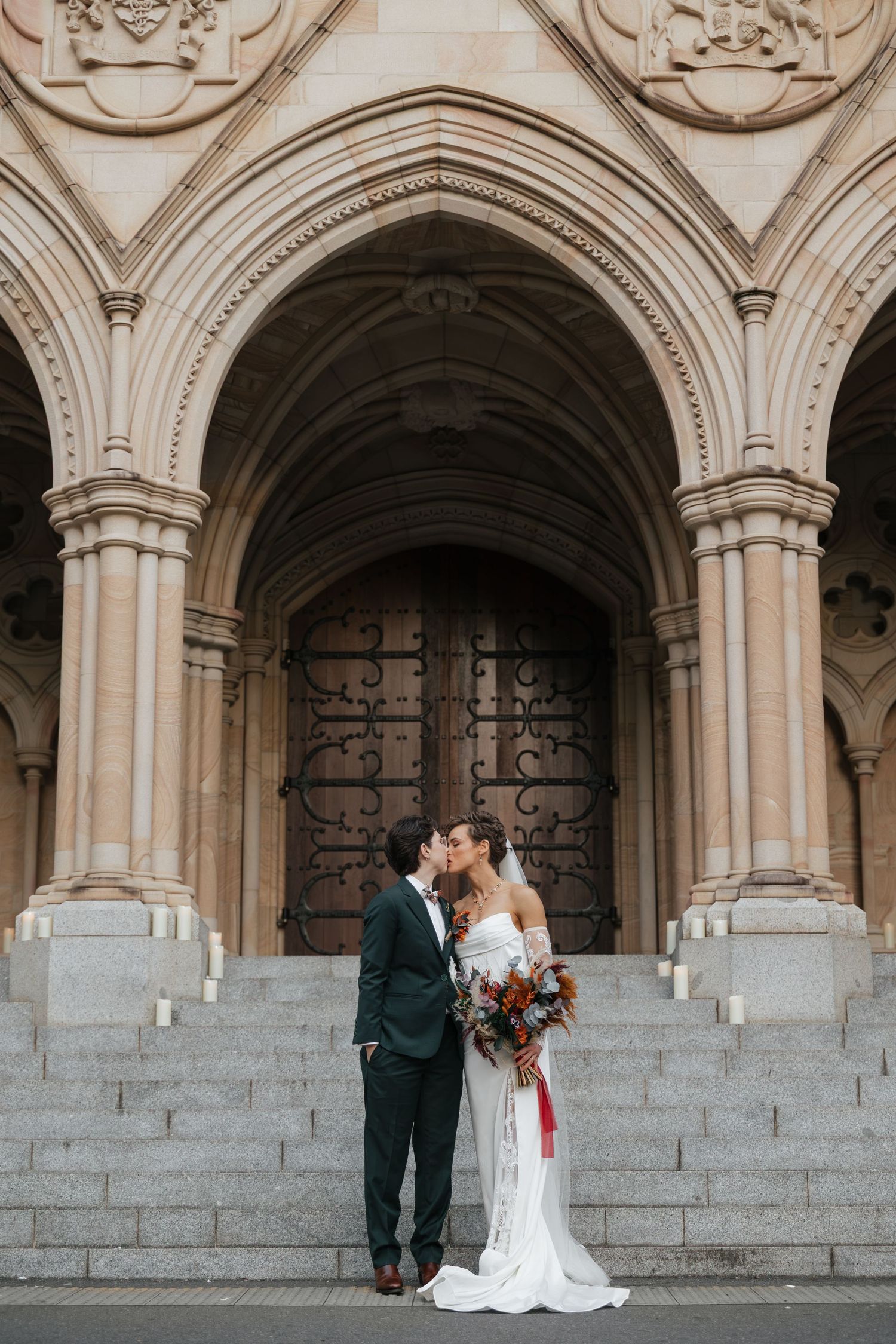Bride and groom kissing on church steps under ornate stone arches, bride holding colorful bouquet.