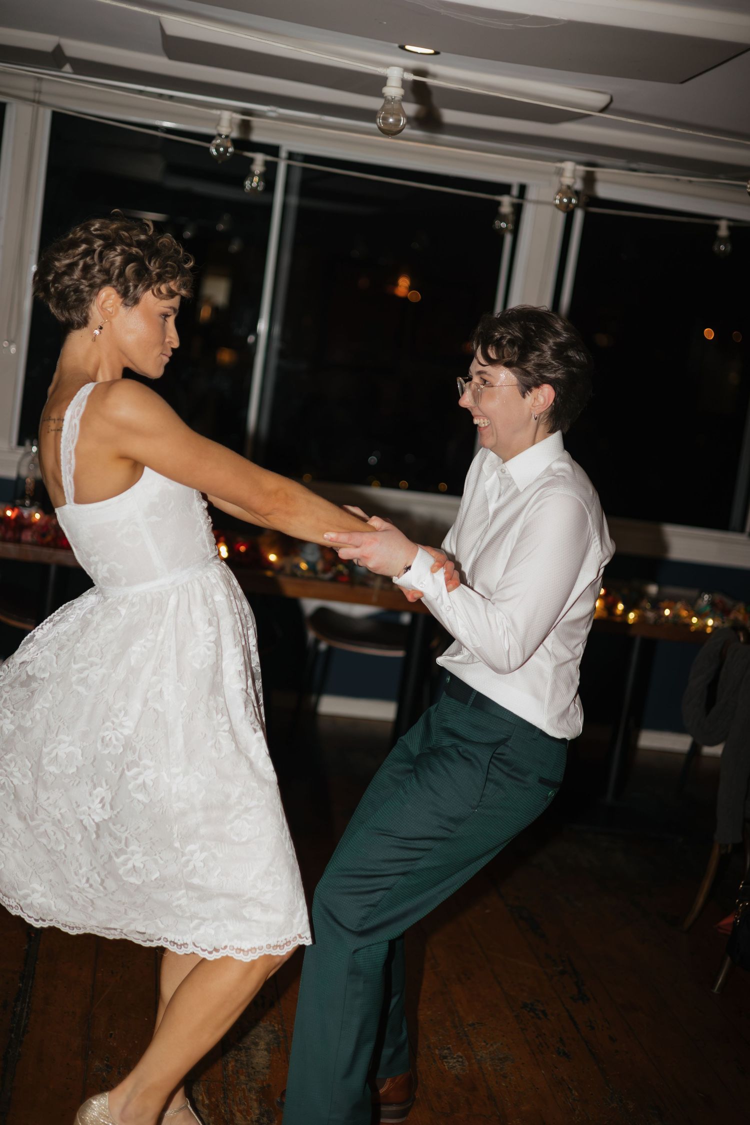 Two brides share a joyful first dance at their wedding reception, one in a white lace dress and the other in green trousers.