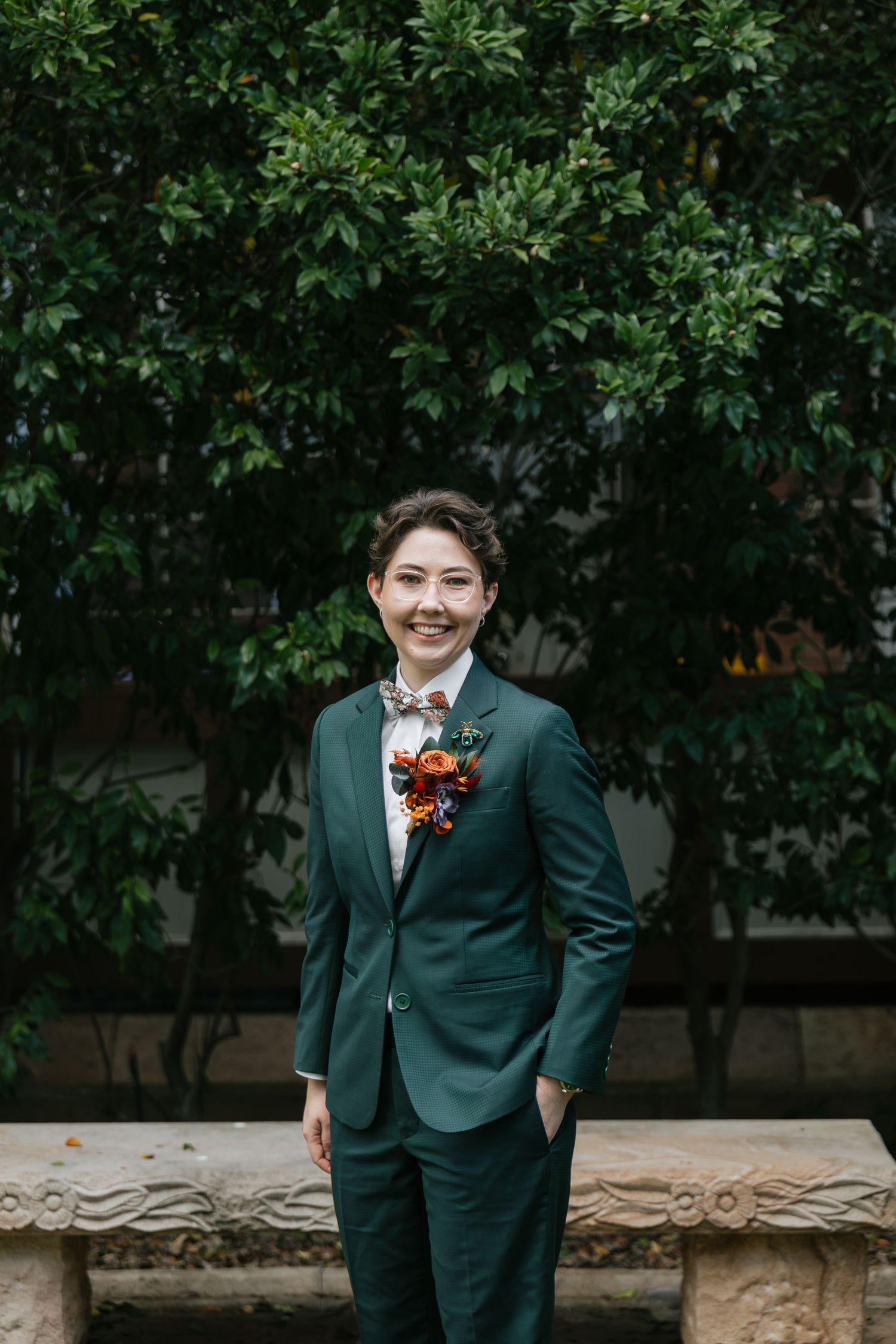 Smiling person in green suit with floral boutonniere poses outdoors against lush green hedge backdrop.