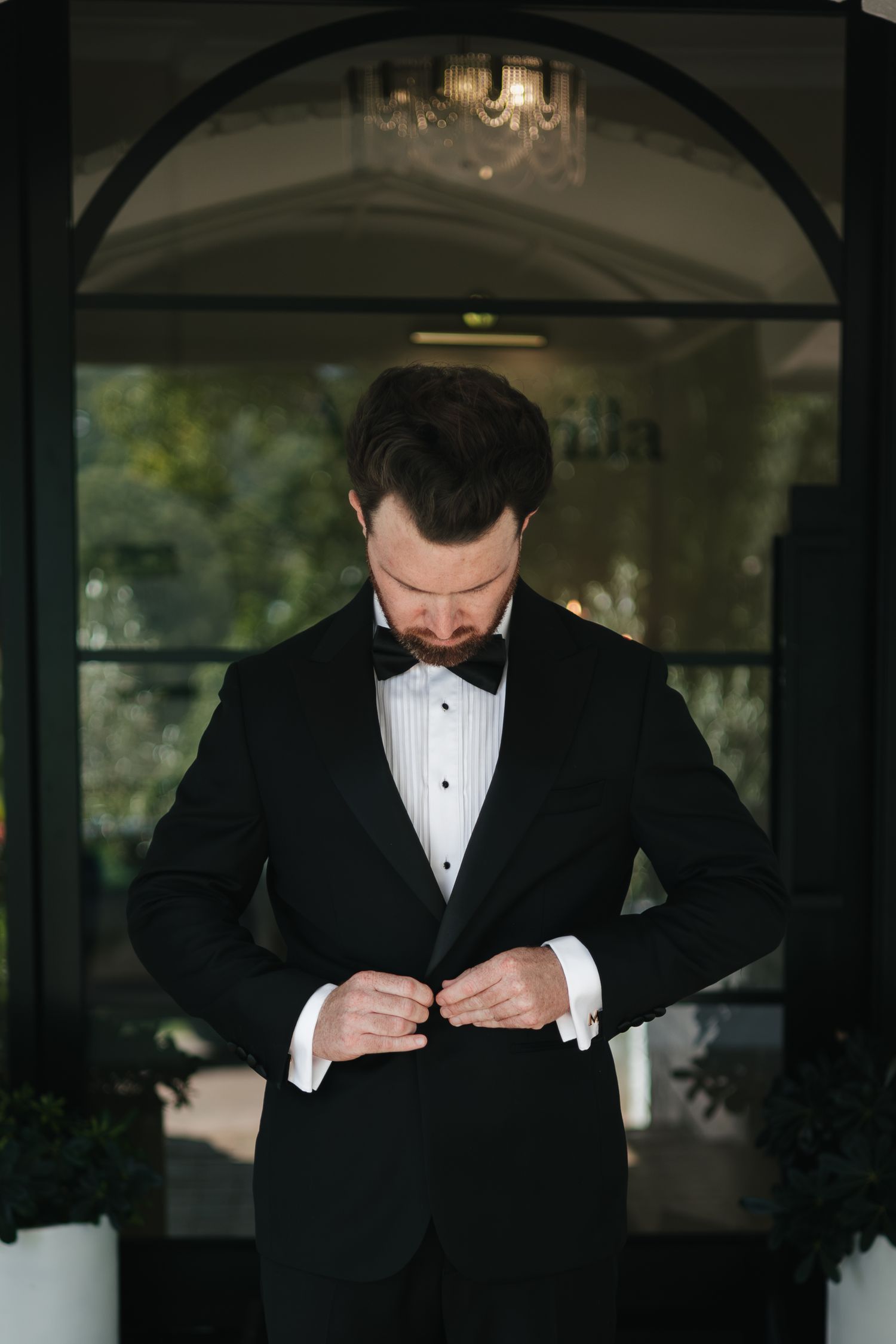 Groom in black tuxedo with bow tie buttoning jacket under elegant arched entrance with chandelier.