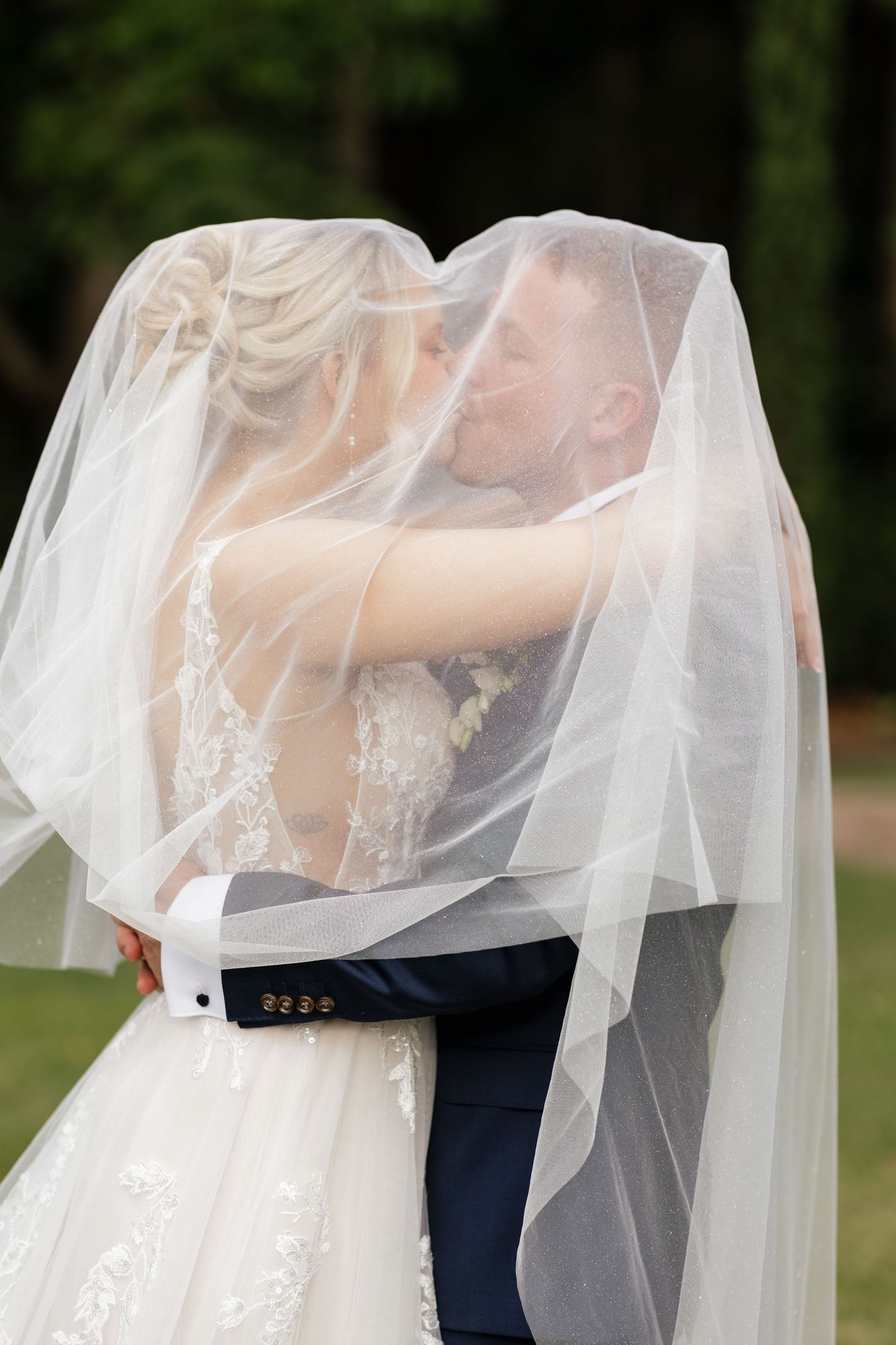 Bride and groom sharing a romantic kiss under a flowing white veil outdoors on their wedding day.