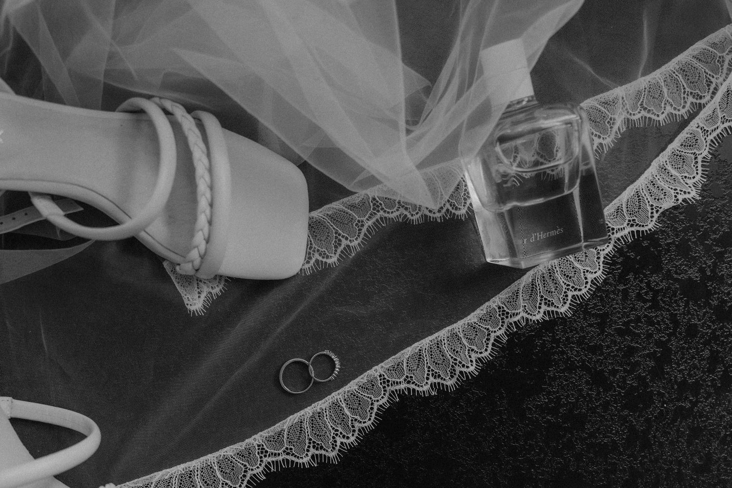 Black and white flat lay of wedding rings, bridal veil, heels, and perfume bottle on dark surface.