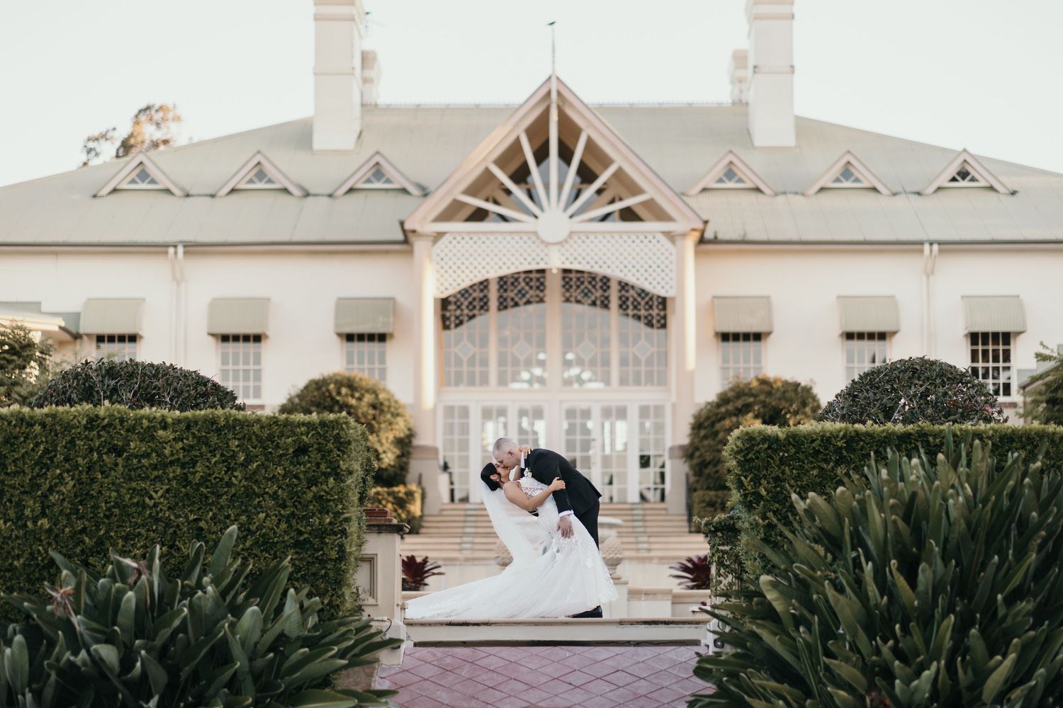 Bride and groom share a romantic dip kiss in a manicured garden in front of an elegant white wedding venue.