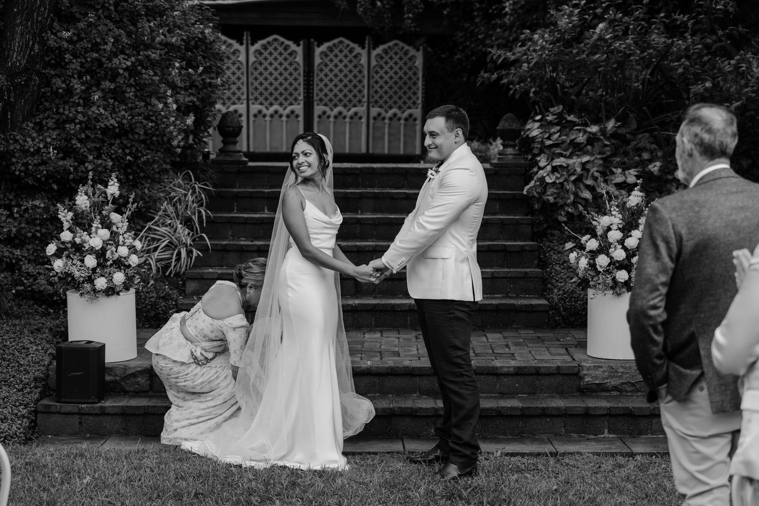 Black and white photo of a bride and groom holding hands during an outdoor wedding ceremony beside floral arrangements.
