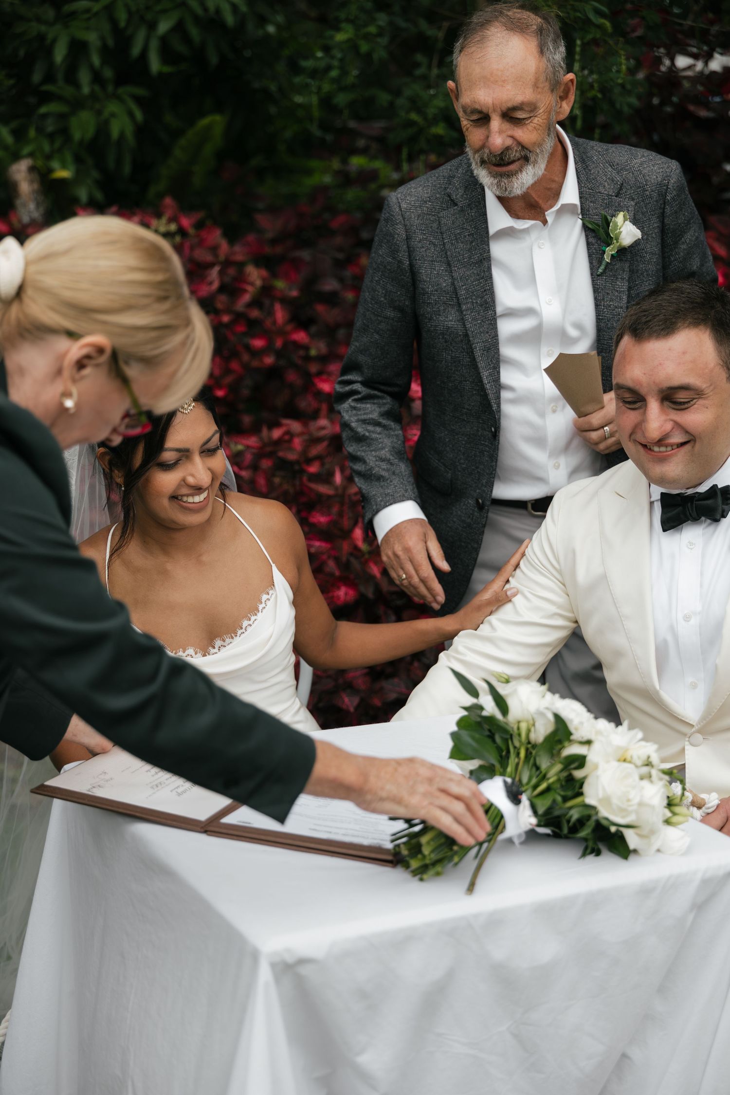 Bride and groom signing wedding register at outdoor ceremony table with officiant and witness present.
