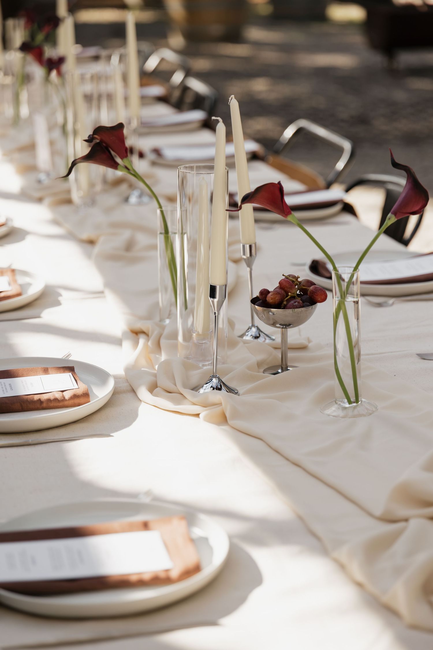 Elegant outdoor wedding table with dark calla lilies, white candles, strawberries, and copper accents on cream linen.