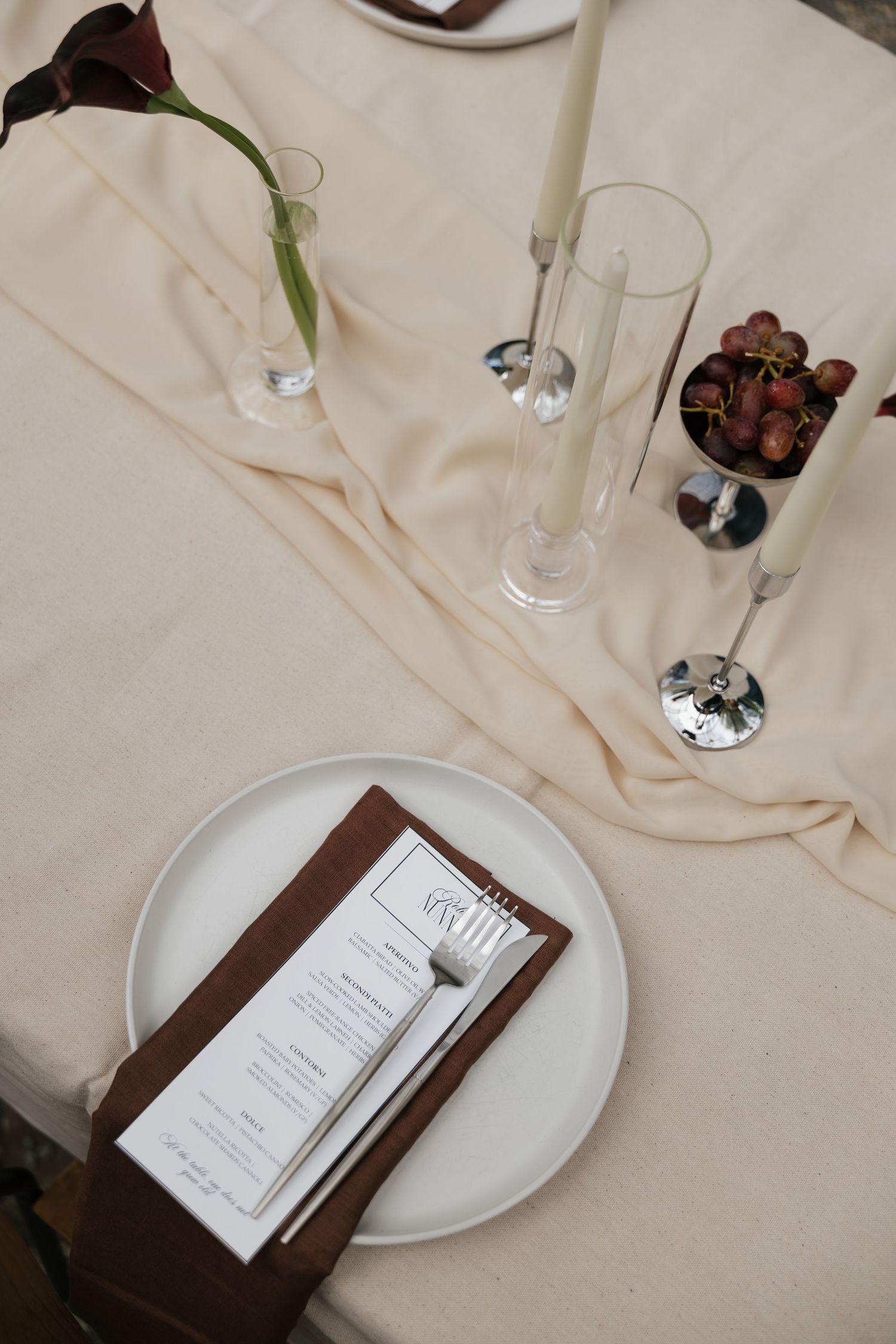 Elegant wedding table setting with cream linen, taper candles, menu card on white plate, and dark red calla lily centerpiece.