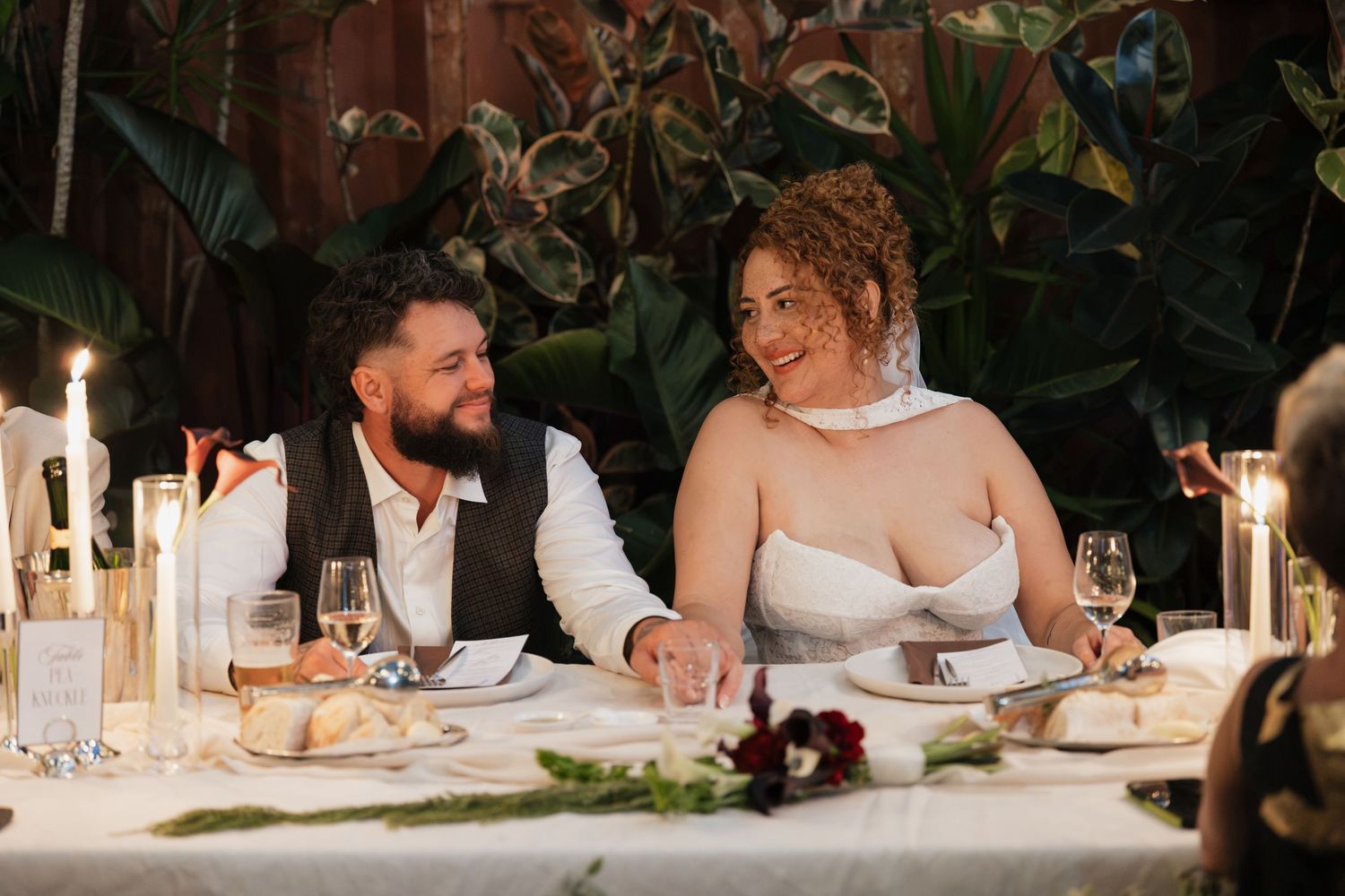 Bride and groom smiling together at wedding reception table with candles and greenery backdrop.