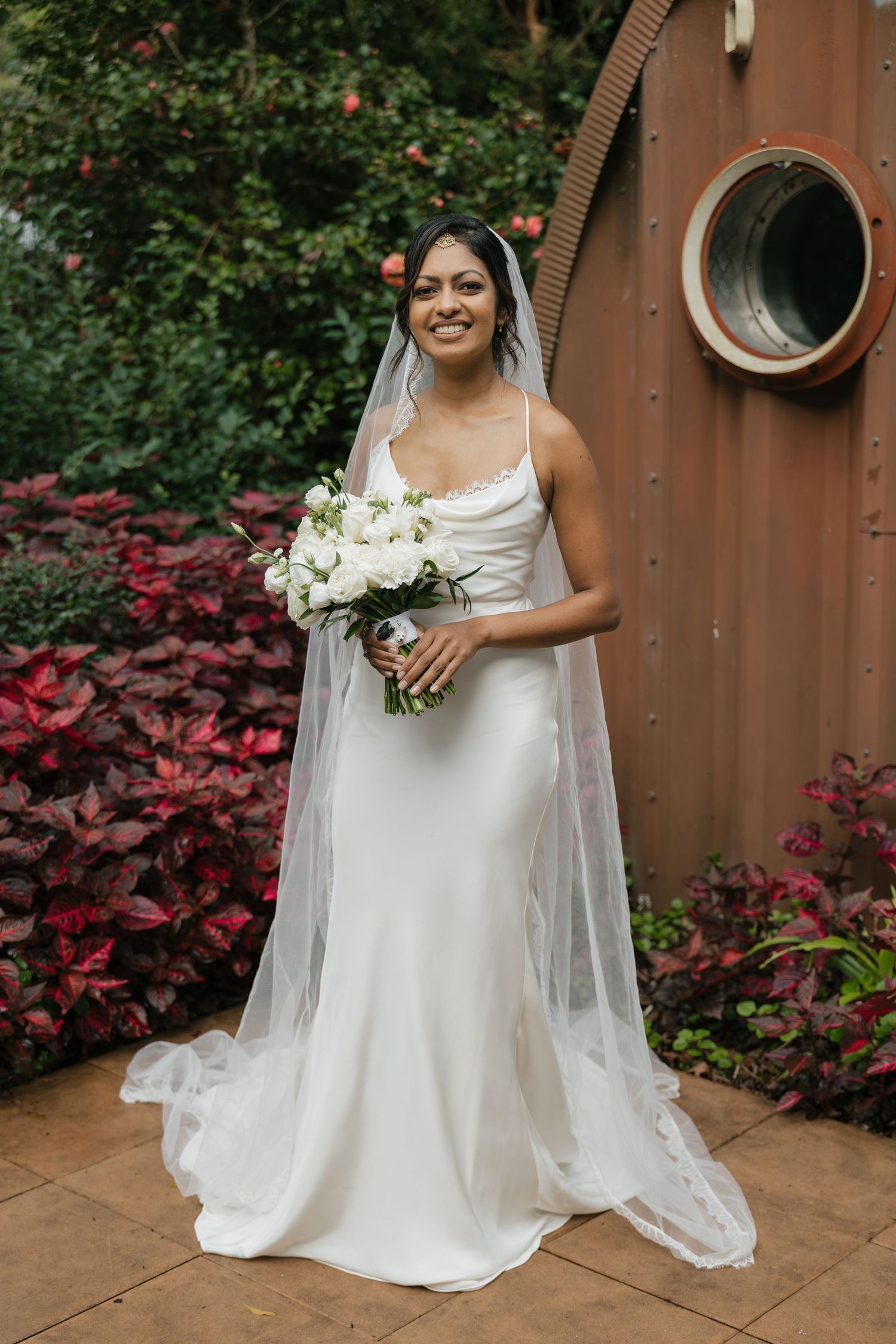 Smiling bride in a white satin gown and long veil holds a white bouquet, standing in a lush garden setting.