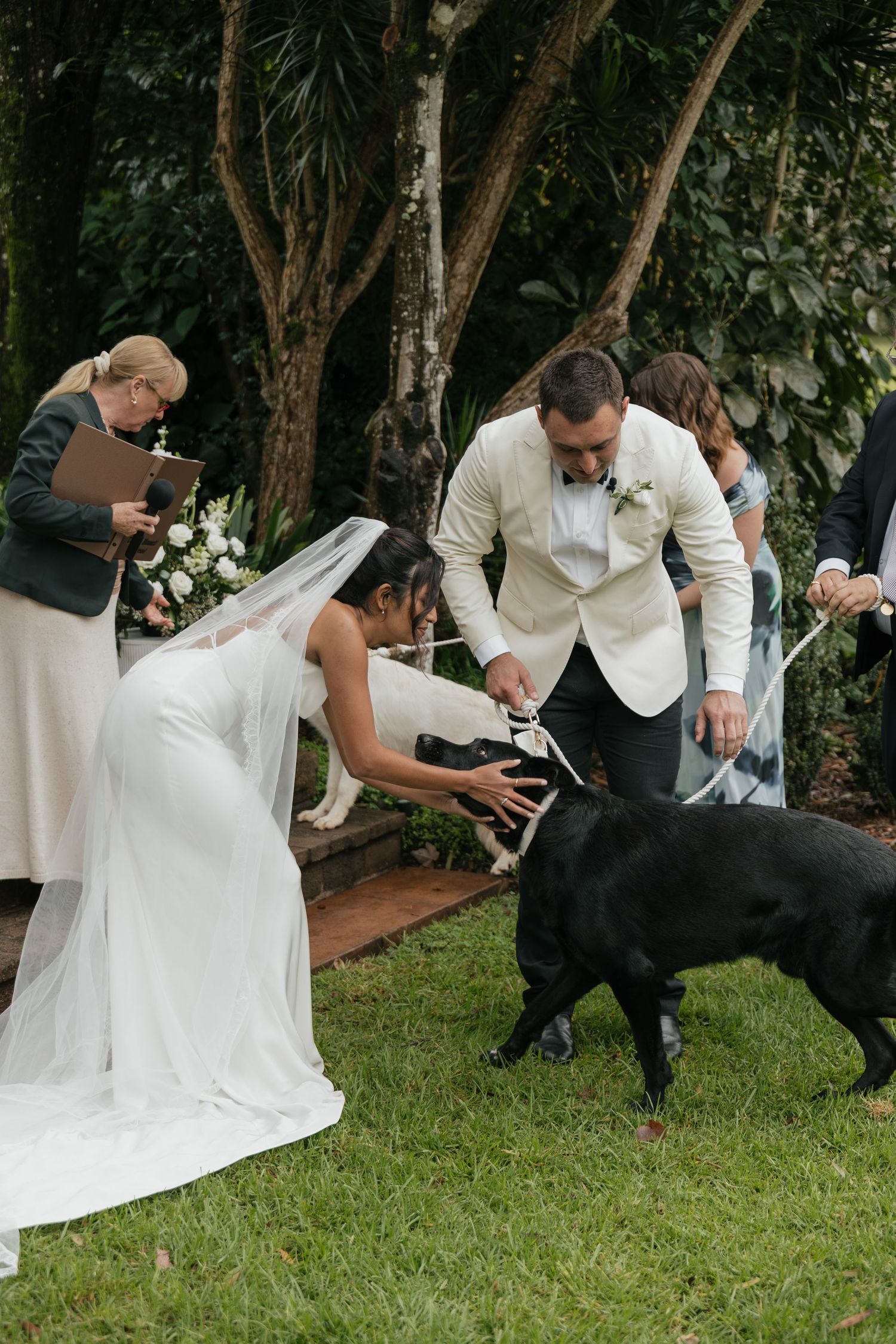 Bride and groom petting a black Labrador dog during an outdoor wedding ceremony surrounded by lush greenery.