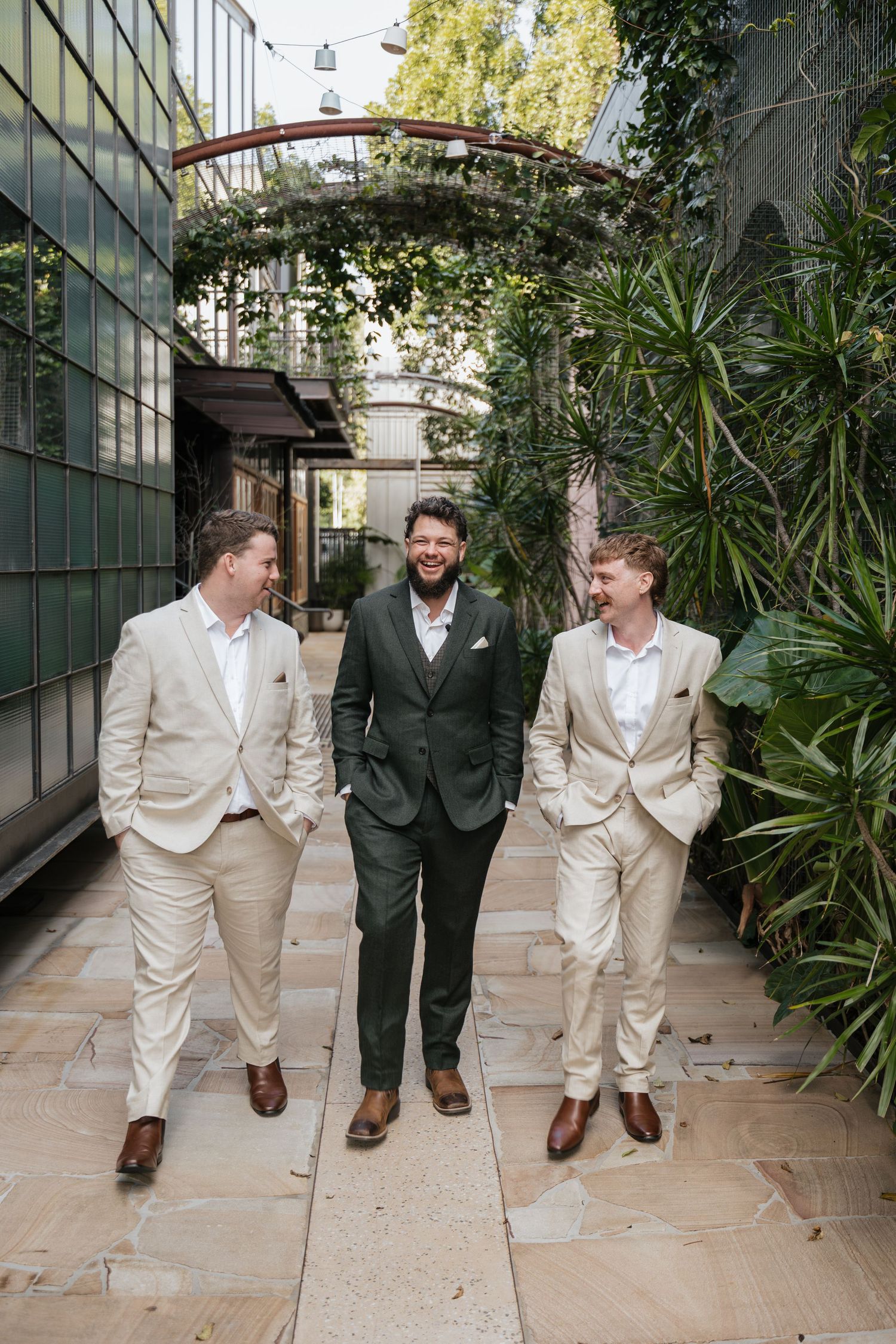Three well-dressed groomsmen walking through a lush garden courtyard, two in beige suits and one in a dark green suit.