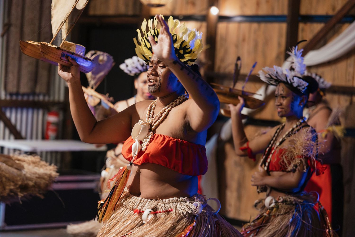 Traditional Pacific Islander dancers in grass skirts and red tops perform a cultural dance ceremony on stage.