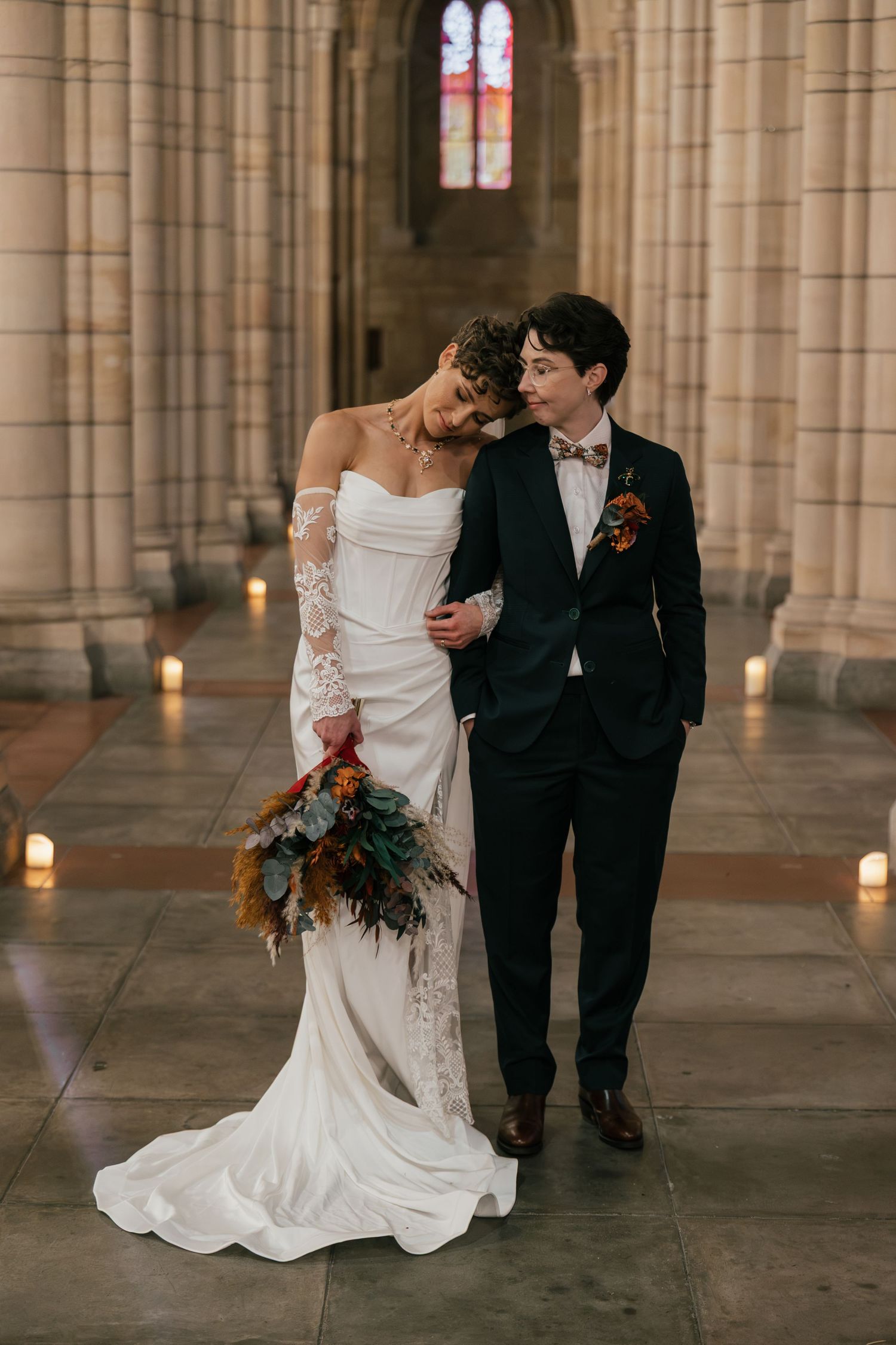 Two brides pose in a candlelit cathedral, one in a white lace gown holding a bouquet, the other in a black suit with boutonniere.
