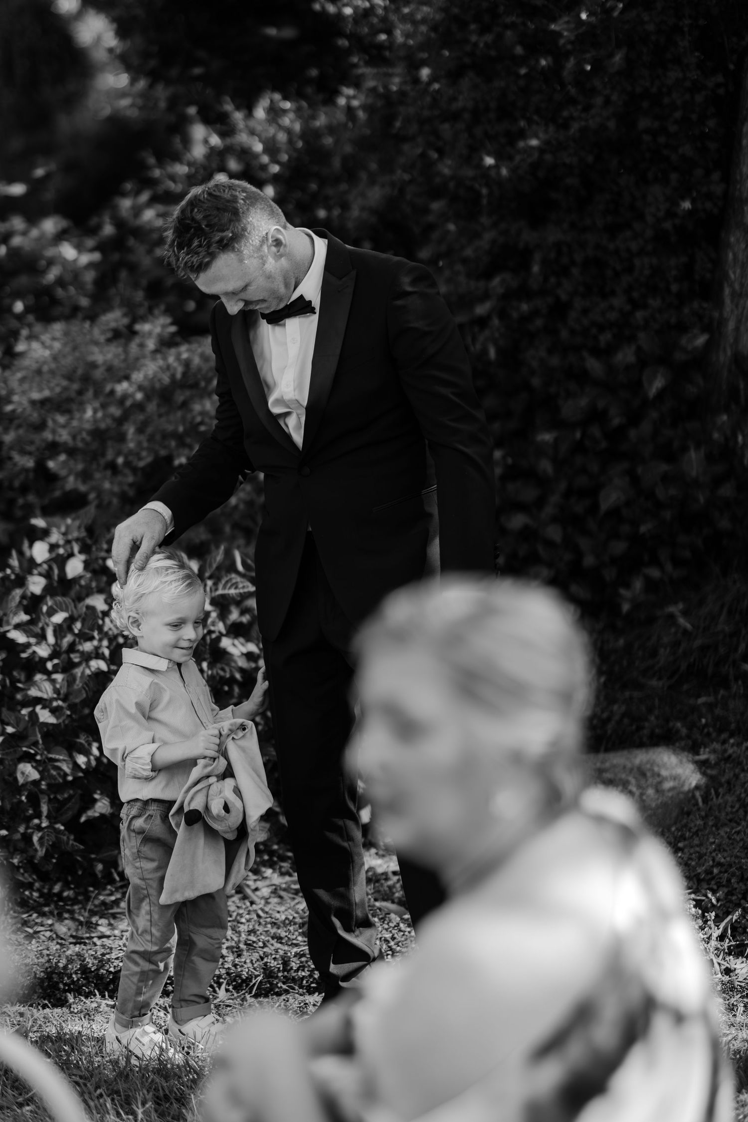 Black and white photo of a groom in tuxedo interacting with a young child at an outdoor wedding reception.