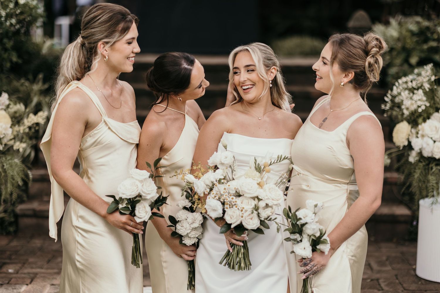 Bride in white strapless gown with three bridesmaids in cream satin dresses holding white floral bouquets outdoors.