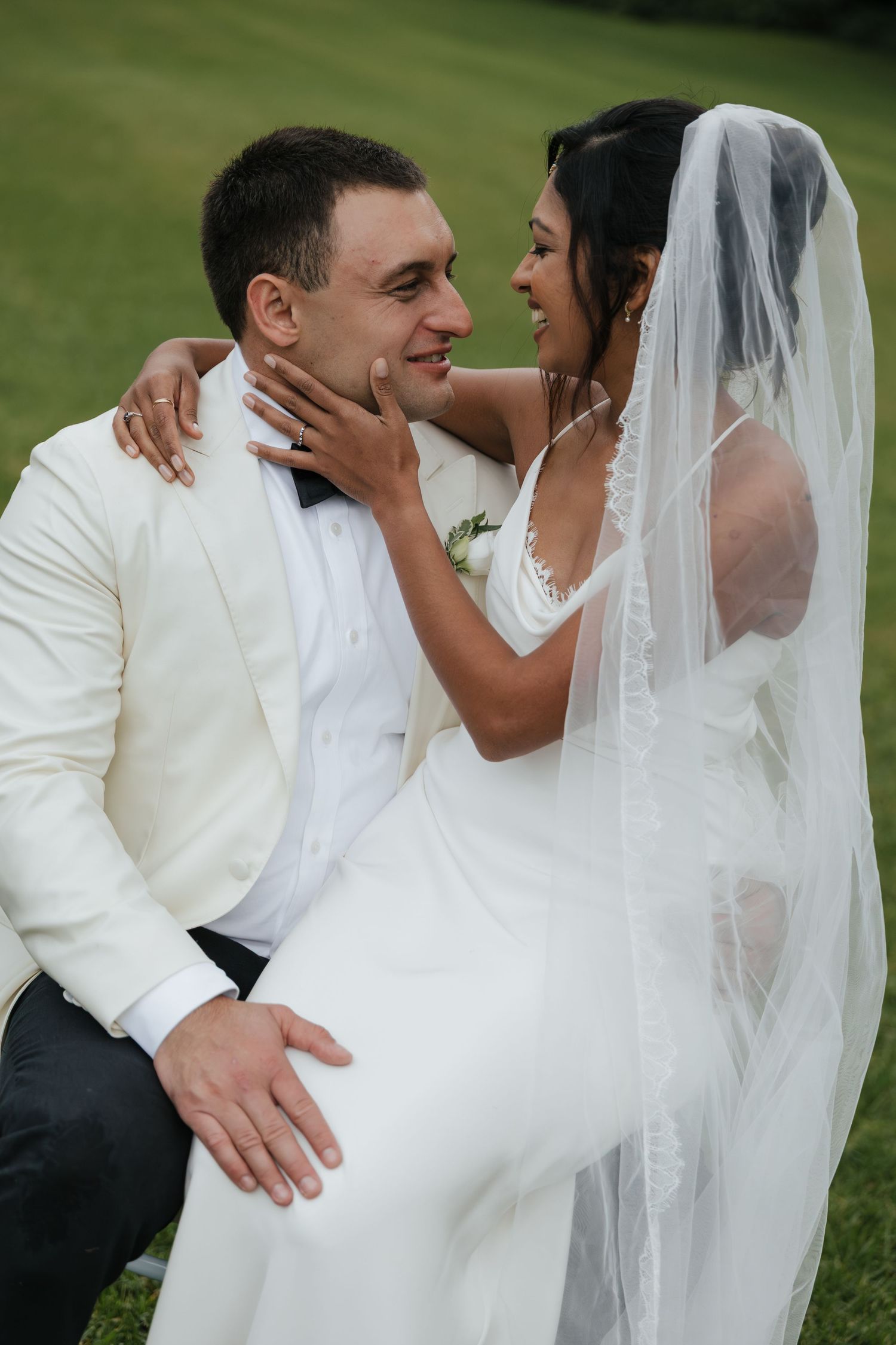 A bride in a white gown and veil tenderly holds a groom's face as they share a romantic moment outdoors.