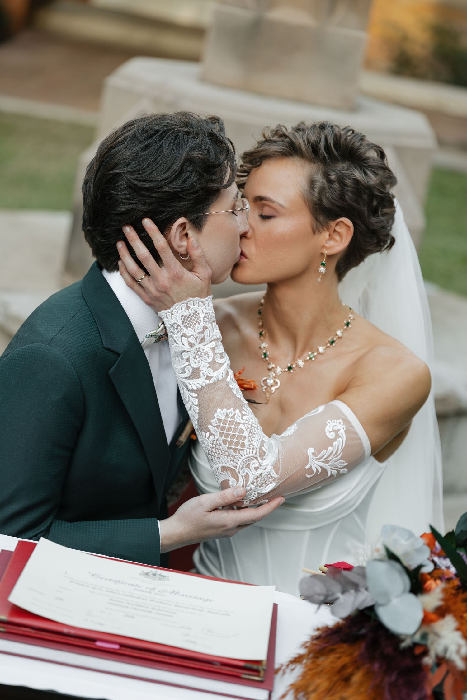Two brides share a kiss at their wedding ceremony signing table, one in a white lace gown, the other in a green suit.