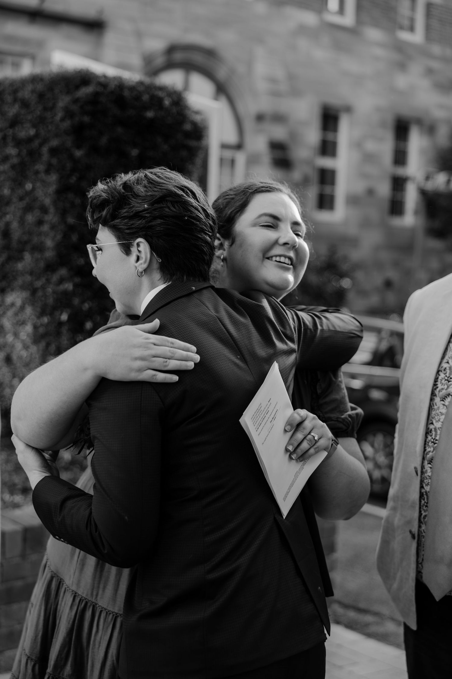 Two women share a joyful hug outdoors near a stone building, captured in black and white photography.