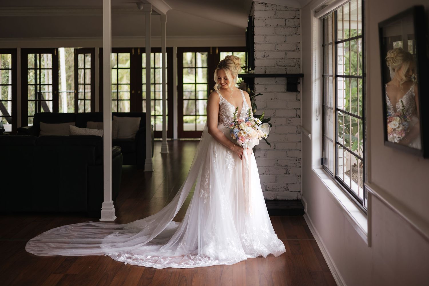 Bride in elegant white wedding gown with long train holding bouquet, standing by window in rustic venue.