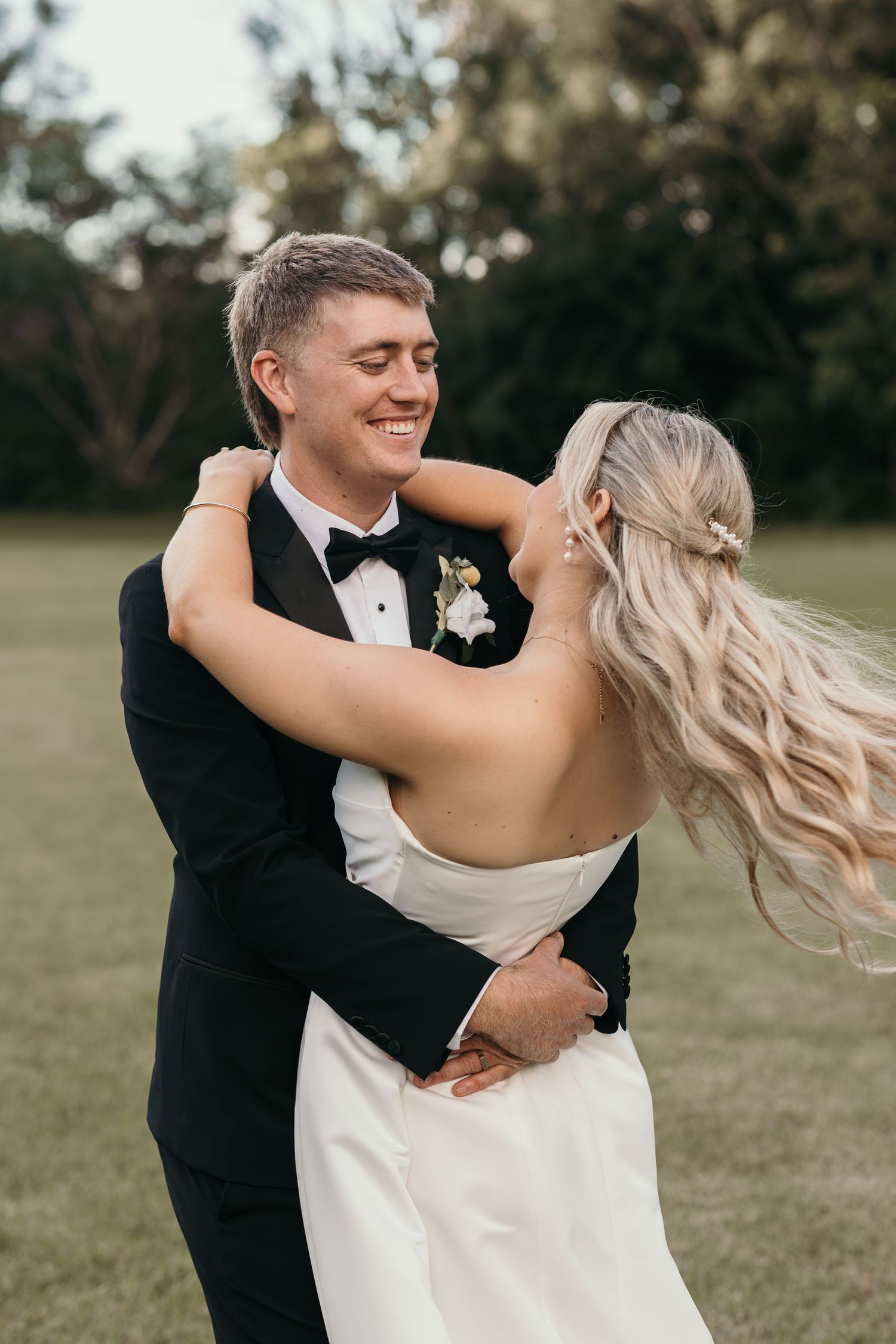 Smiling groom in black tuxedo embraces blonde bride in white wedding dress outdoors on lush green lawn.