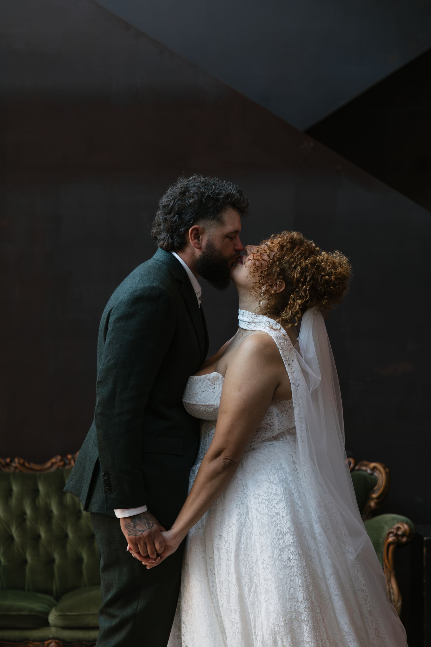 A bride and groom share a romantic kiss while holding hands in an elegant dark-toned venue.